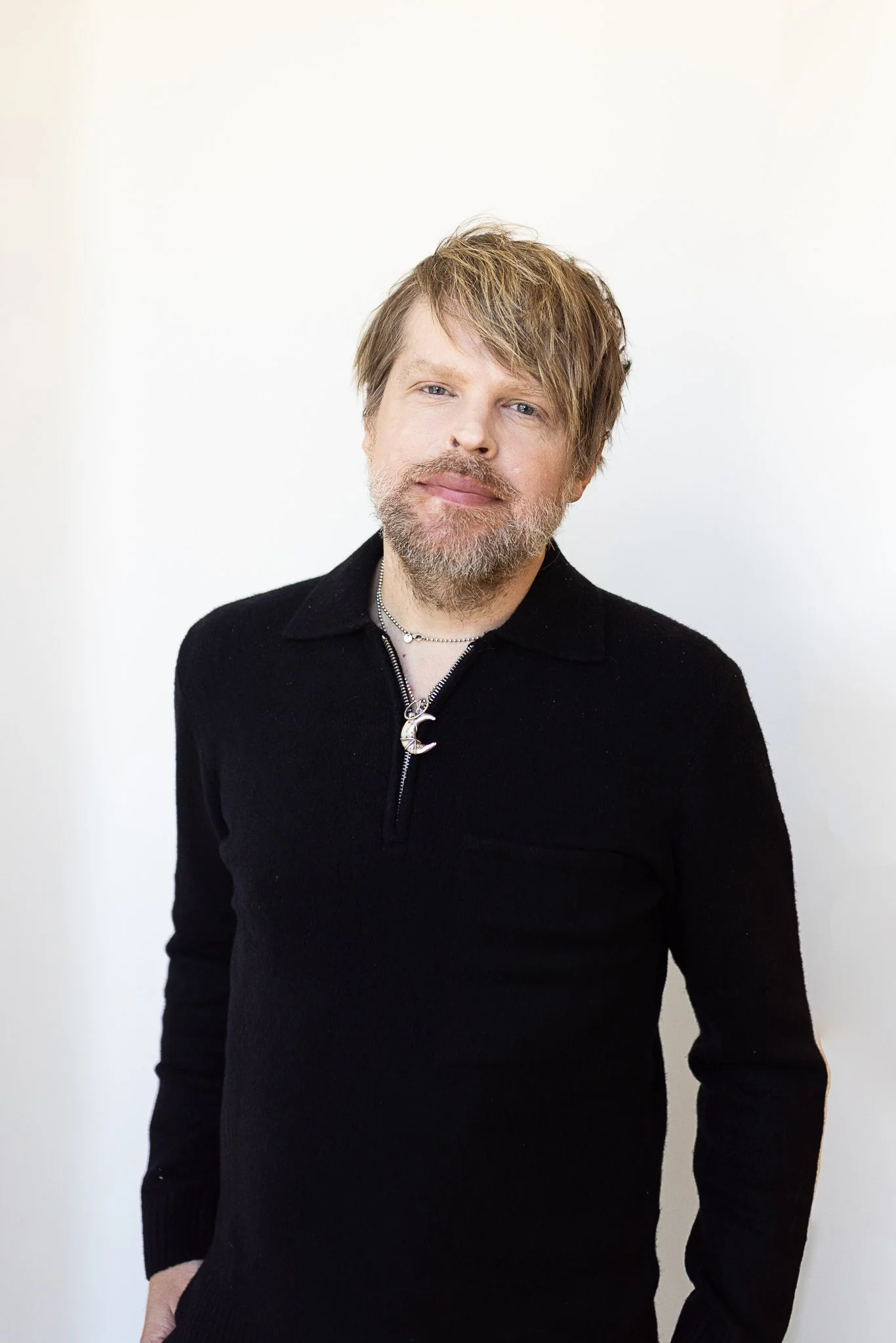 A stylist with light brown, slightly tousled hair and a beard, wearing a black quarter sweater