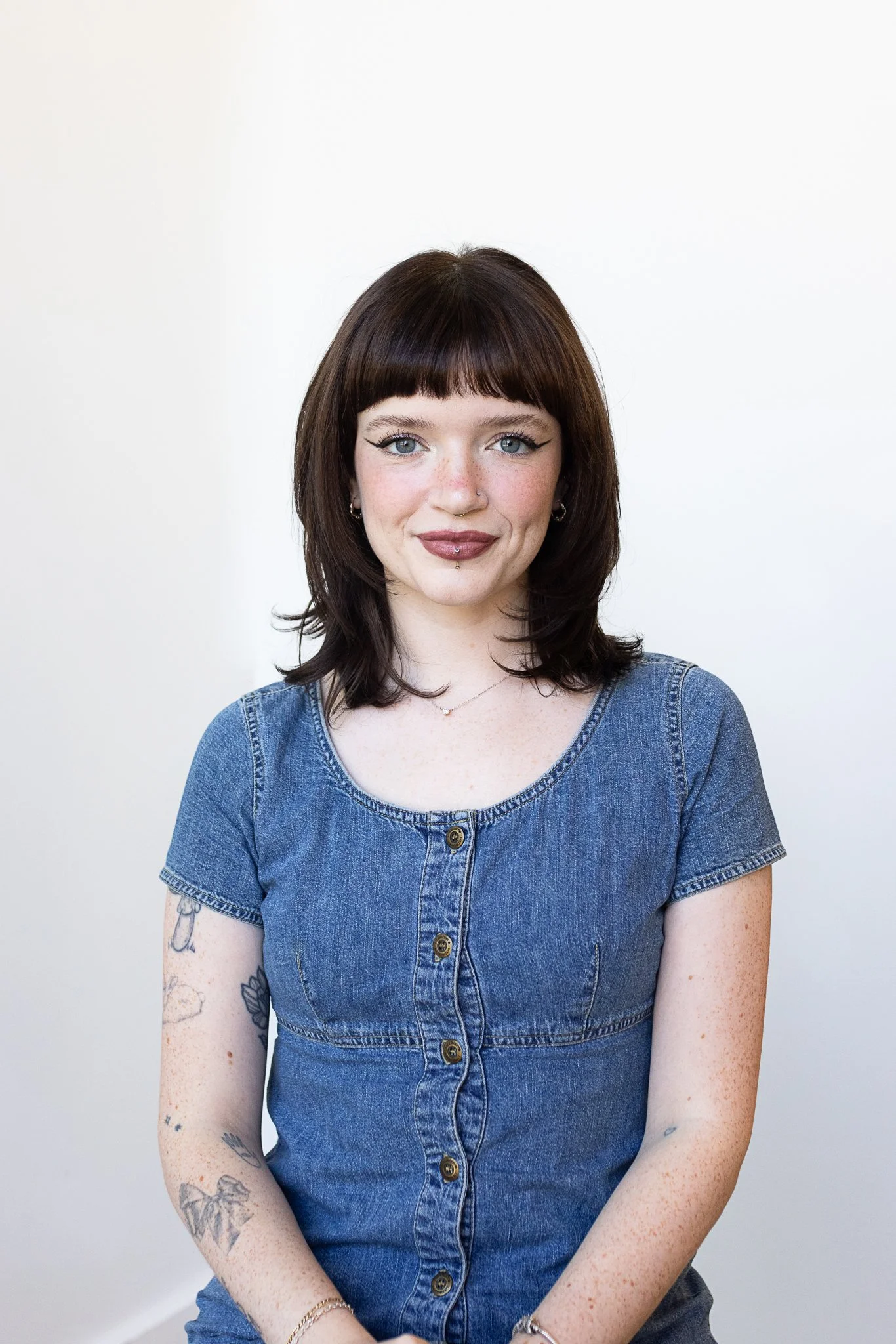 A stylist with shoulder-length dark brown straight hair and blunt bangs, wearing a denim dress