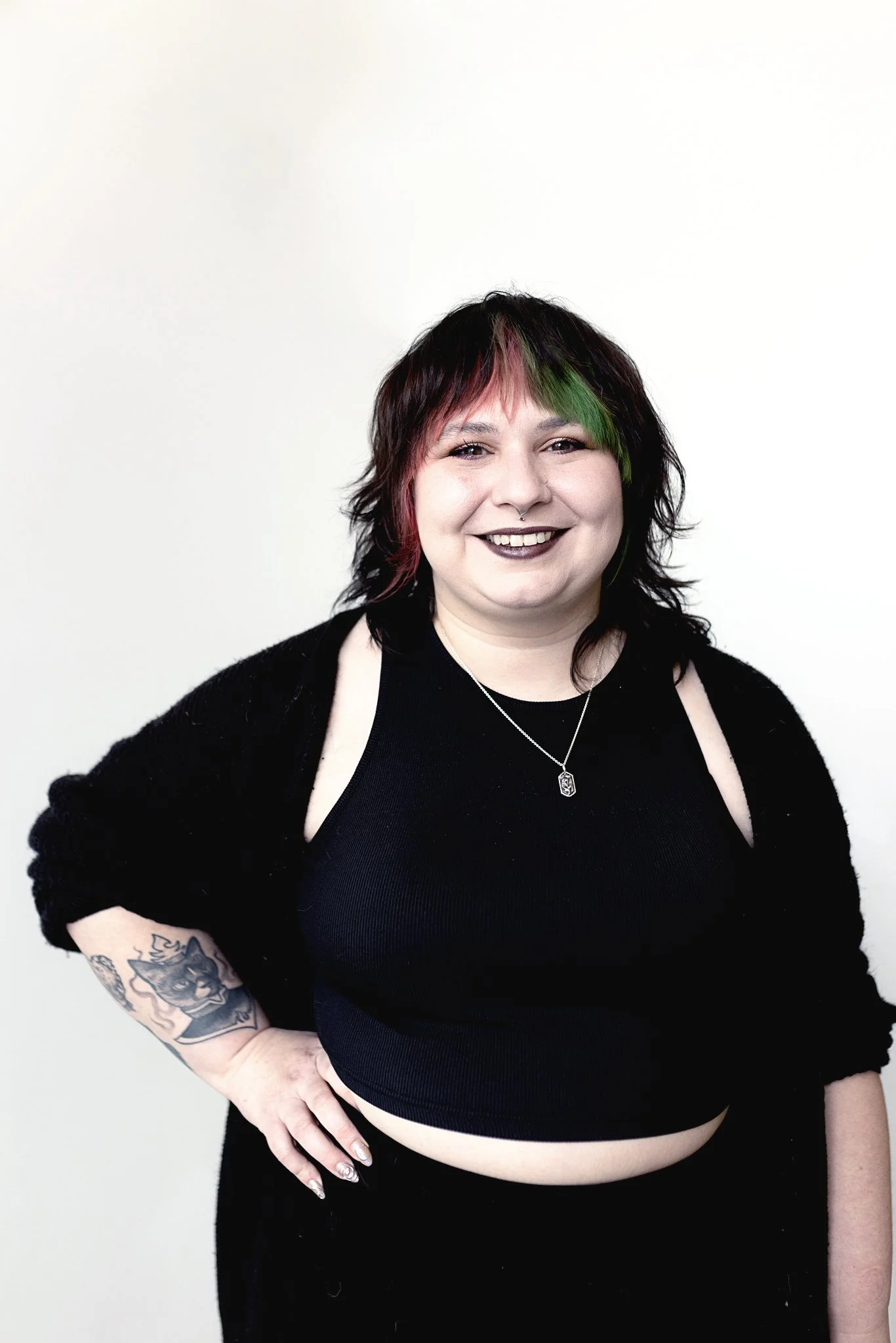 A stylist with shoulder length, red and green-tinted wavy hair and bangs, wearing a black top