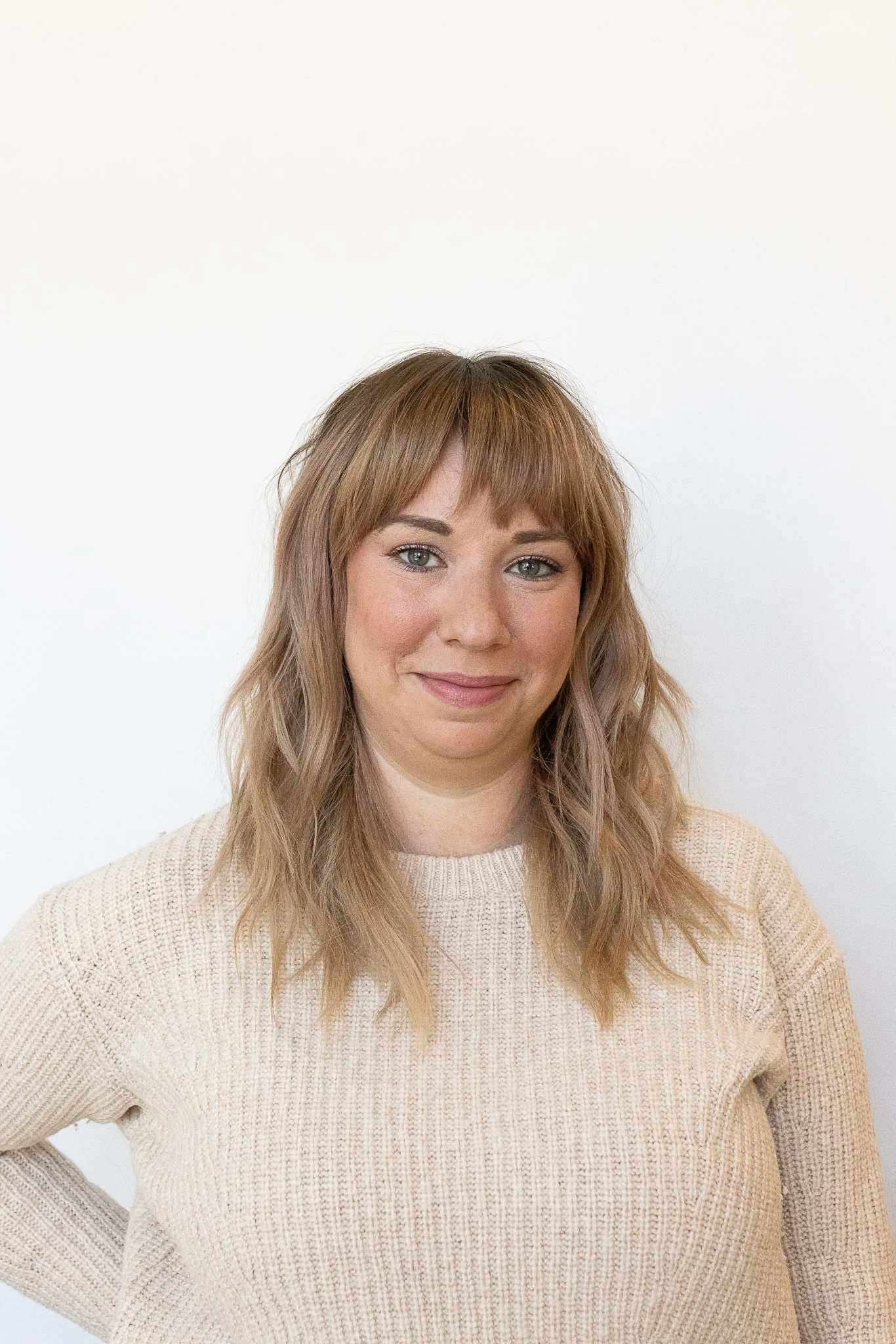 A stylist with shoulder-length wavy light brown hair and bangs, wearing a beige sweater