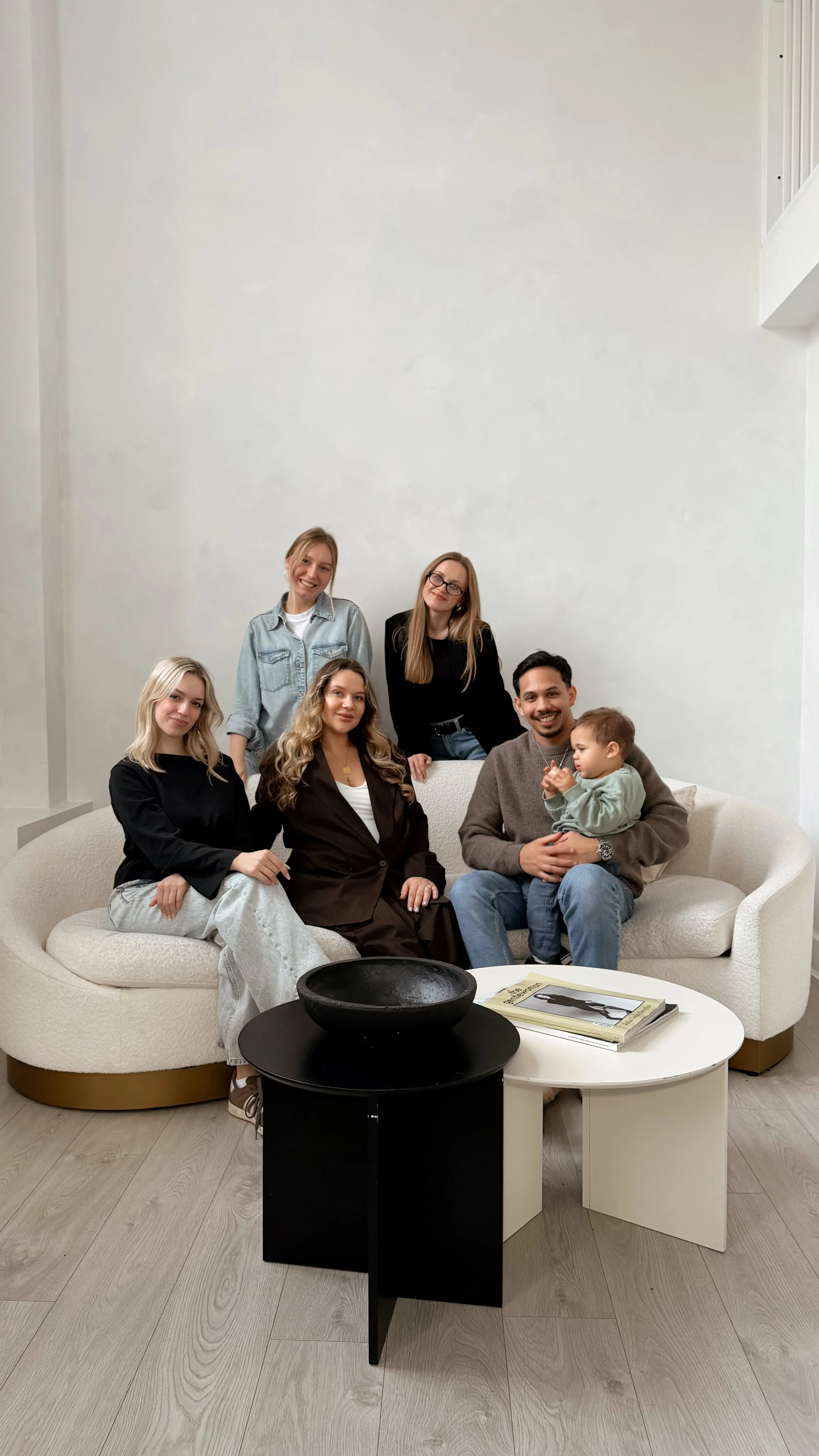 Group of six people, five women and one man, sitting and standing around a white curved sofa in a modern living room. One of the women is holding a small child. A black decorative bowl and a white coffee table with books are in the foreground.