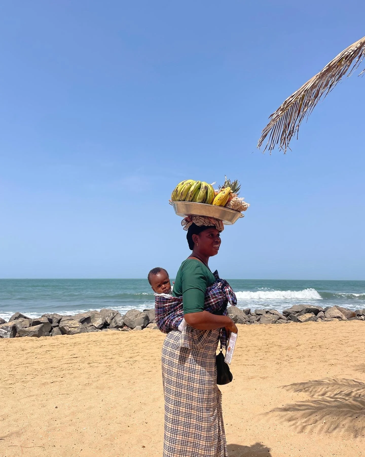 A woman walking on the beach carries a child on her back while balancing a tray of bananas and other fruit on her head. The beach has sand, rocks, and ocean waves under a clear blue sky.