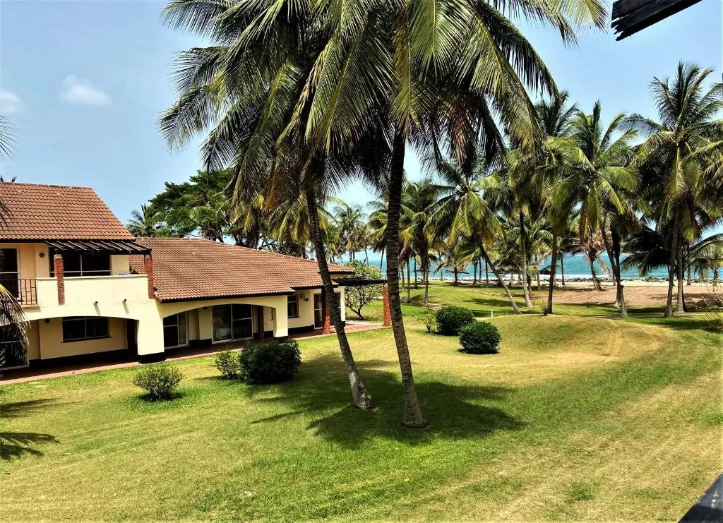 Beachfront villa with red-tiled roof, surrounded by green grass and palm trees, overlooking the beach and ocean.