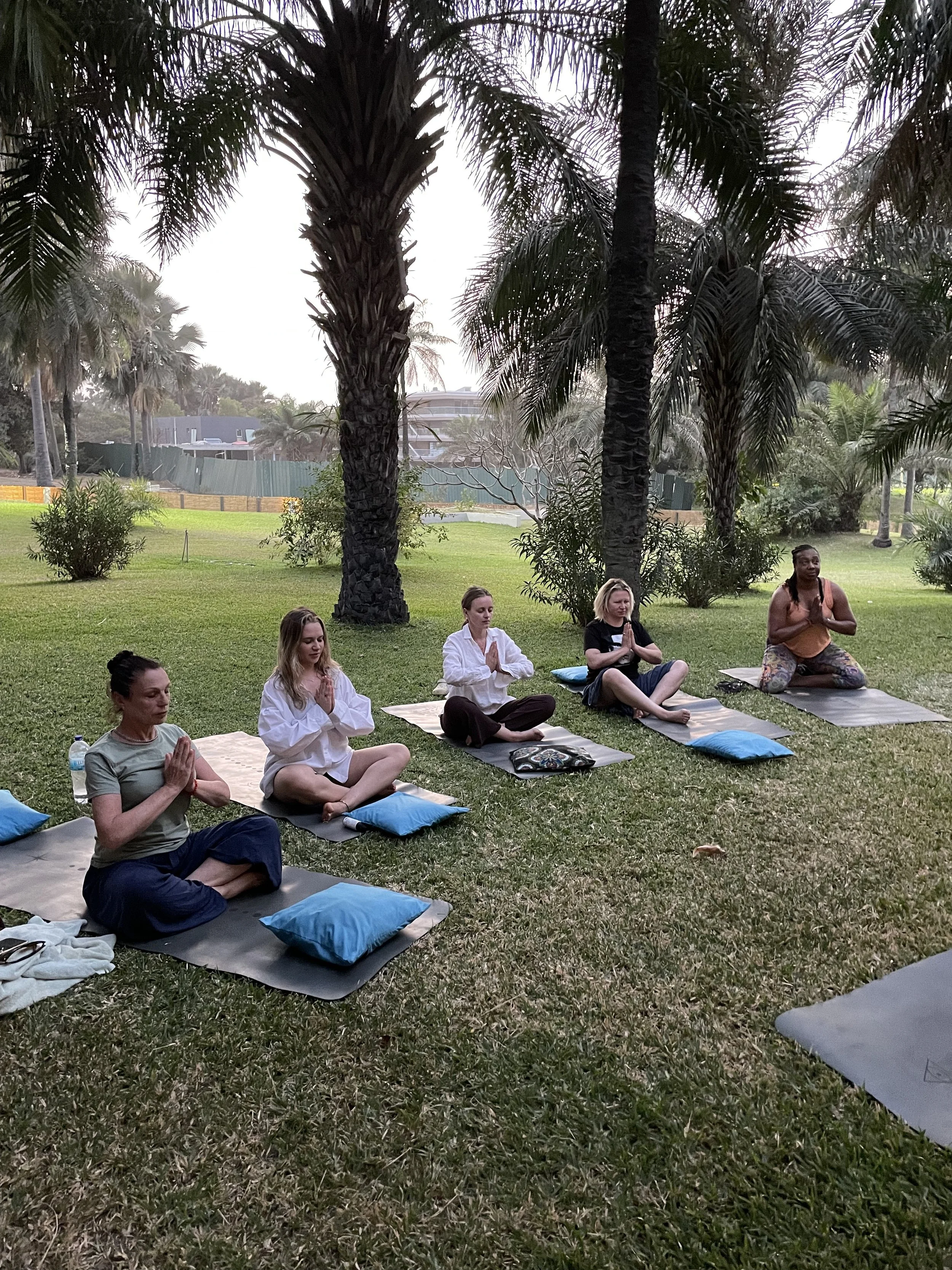A group of five women practicing yoga outdoors in a park, sitting on yoga mats under palm trees with their hands in prayer position.