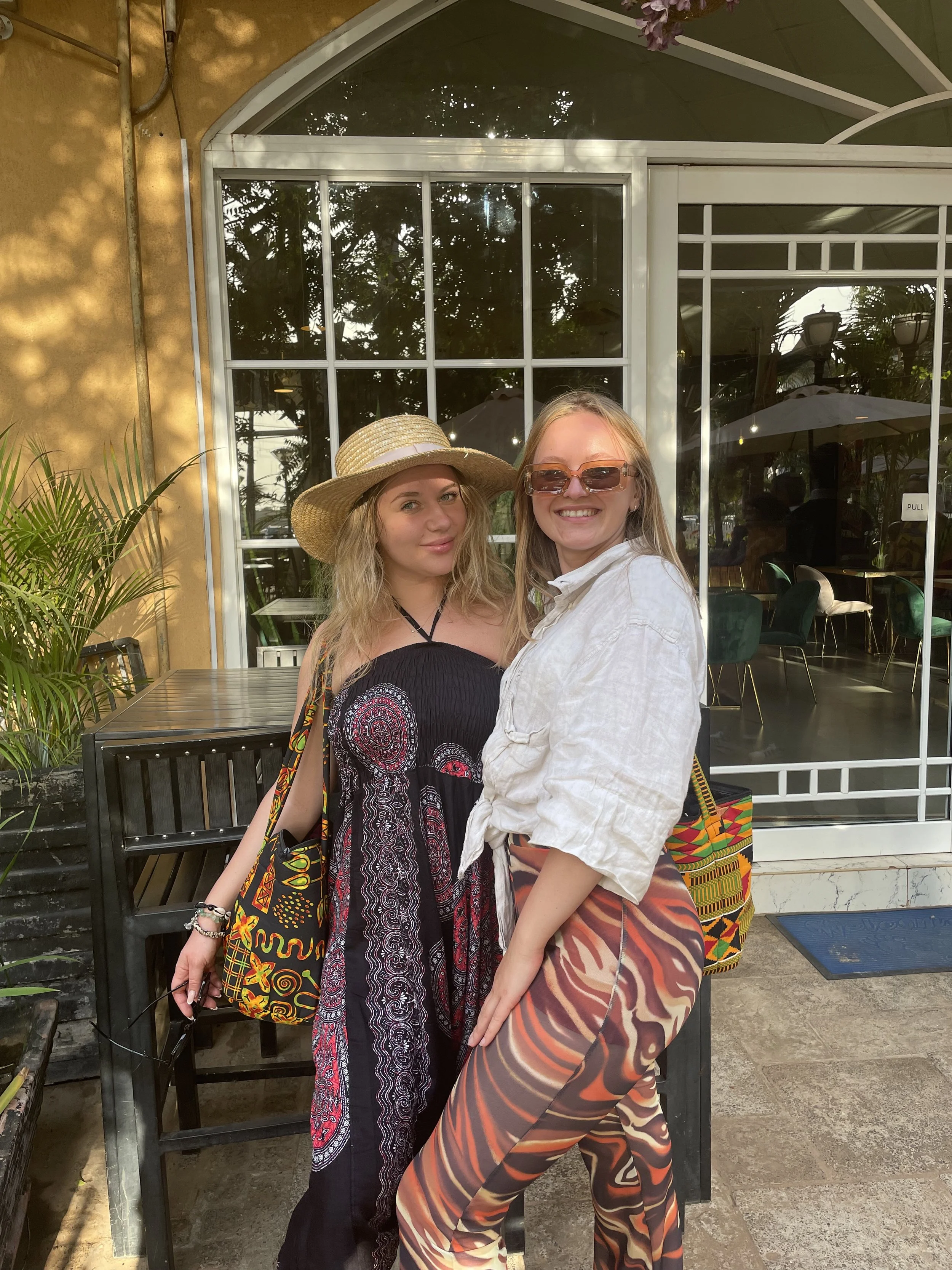 Two women standing outside a restaurant entrance, smiling at the camera. One is wearing a large straw hat and a strapless black dress with colorful patterns, the other has sunglasses, a white shirt, and patterned pants. There are plants and a glass door behind them.