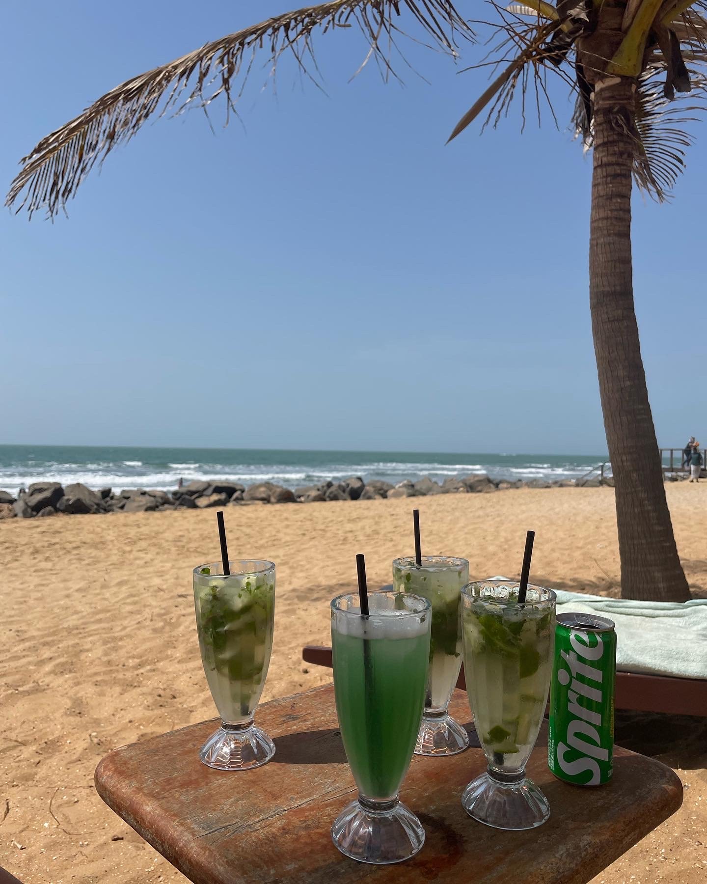 Four glasses of green cocktails with mint and ice on a wooden table at the beach, with a palm tree, sandy shore, rocks, ocean waves, and a clear blue sky in the background, alongside a can of Sprite.