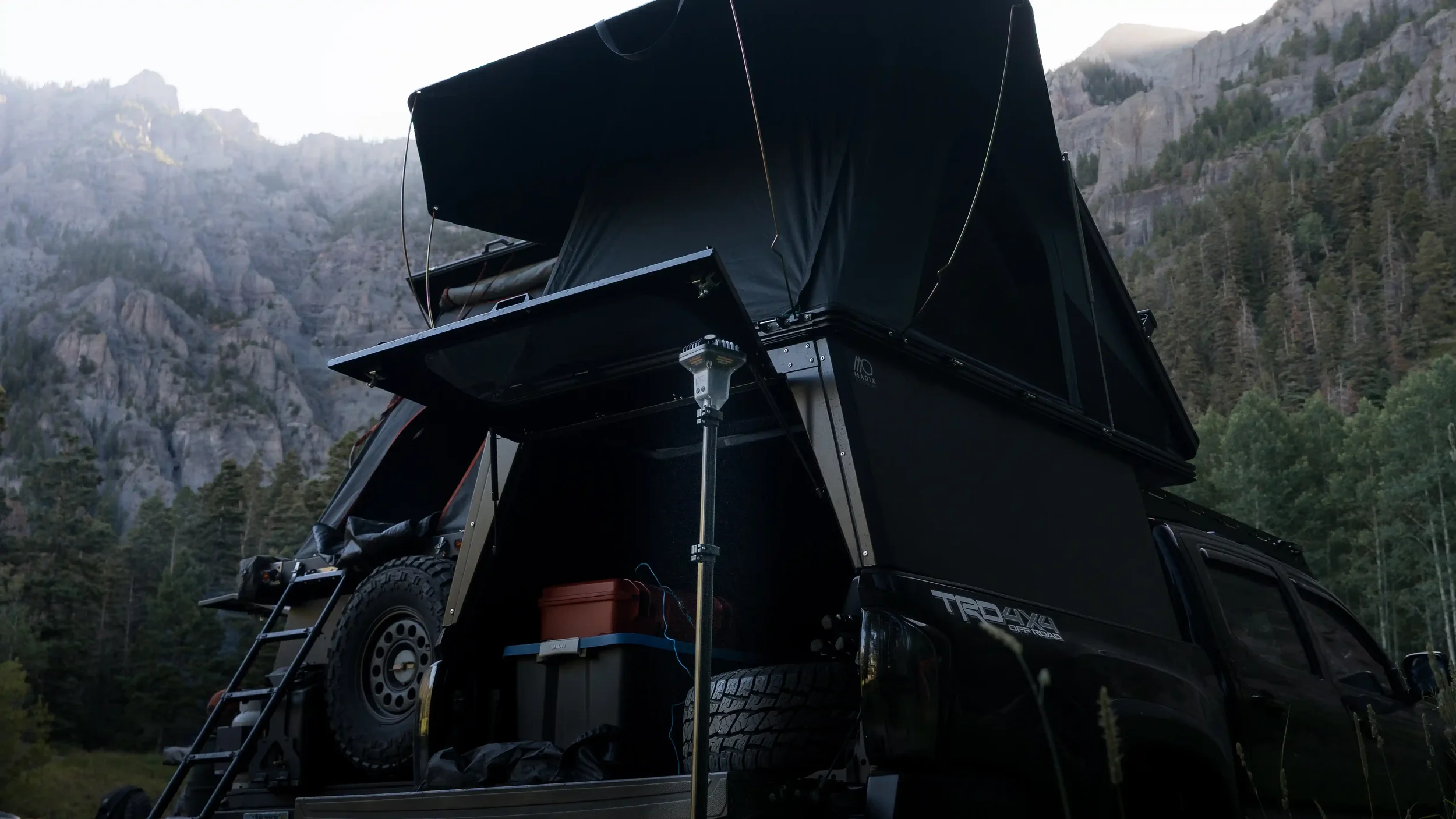 Off-road vehicle with a camper and rooftop tent set up, parked in a mountainous area with trees and rocky cliffs in the background.