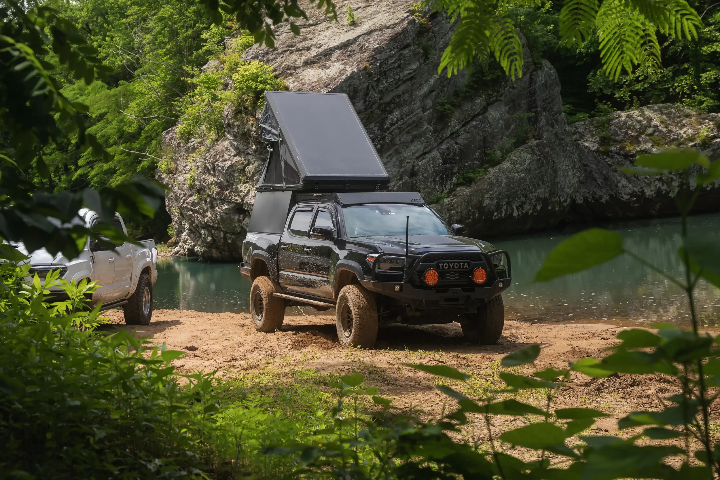 Toyota Tacoma build with pop-up camper parked near a river in the forest.