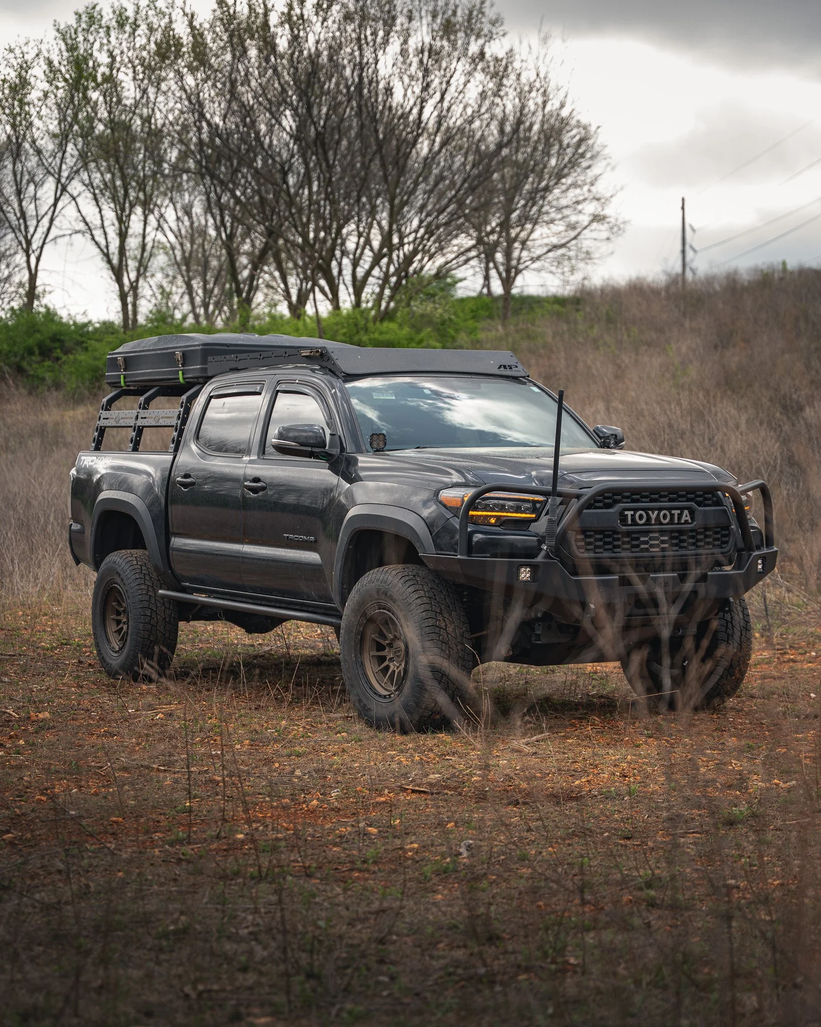 A black Toyota Tacoma pickup truck with off-road tires, a front grille guard bumper, and a rooftop tent parked on a dirt field with trees and overcast sky in the background.