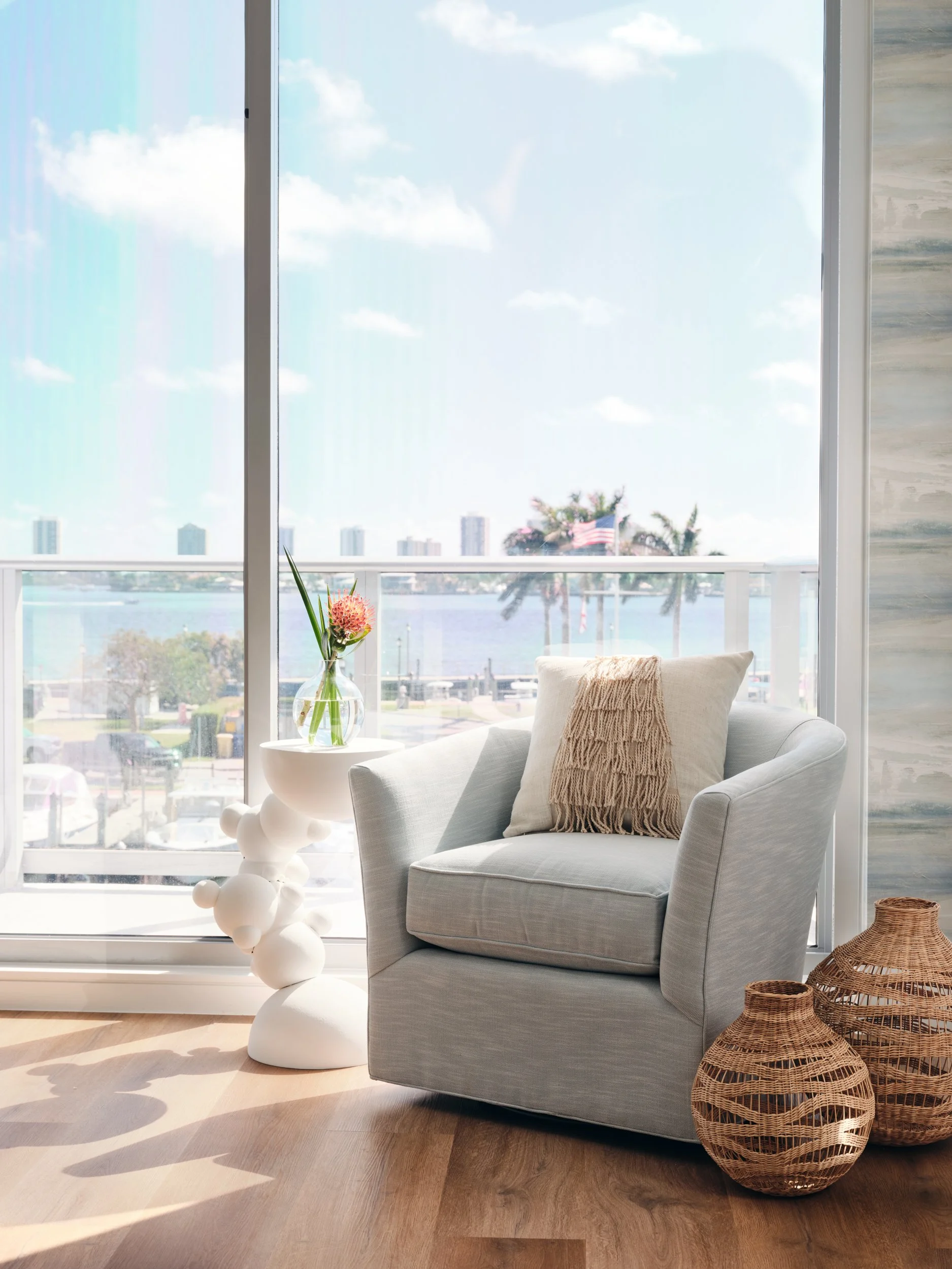 Swivel chair, sculptural white side table and woven vases by the window in a waterfront primary bedroom