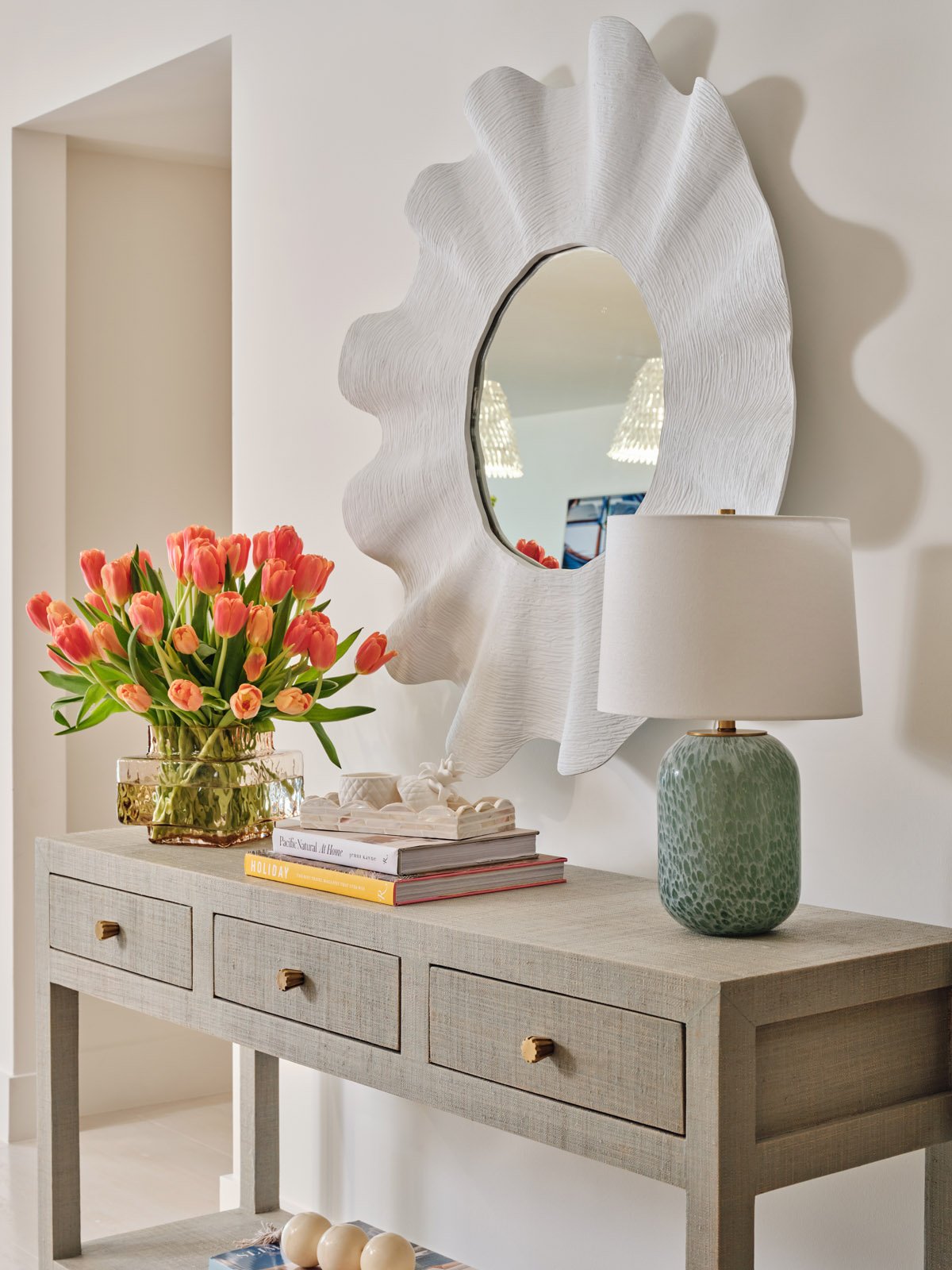 A decorative white textured wall mirror, a vase with pink tulips, a stack of books, and a green textured table lamp on a wooden console table.