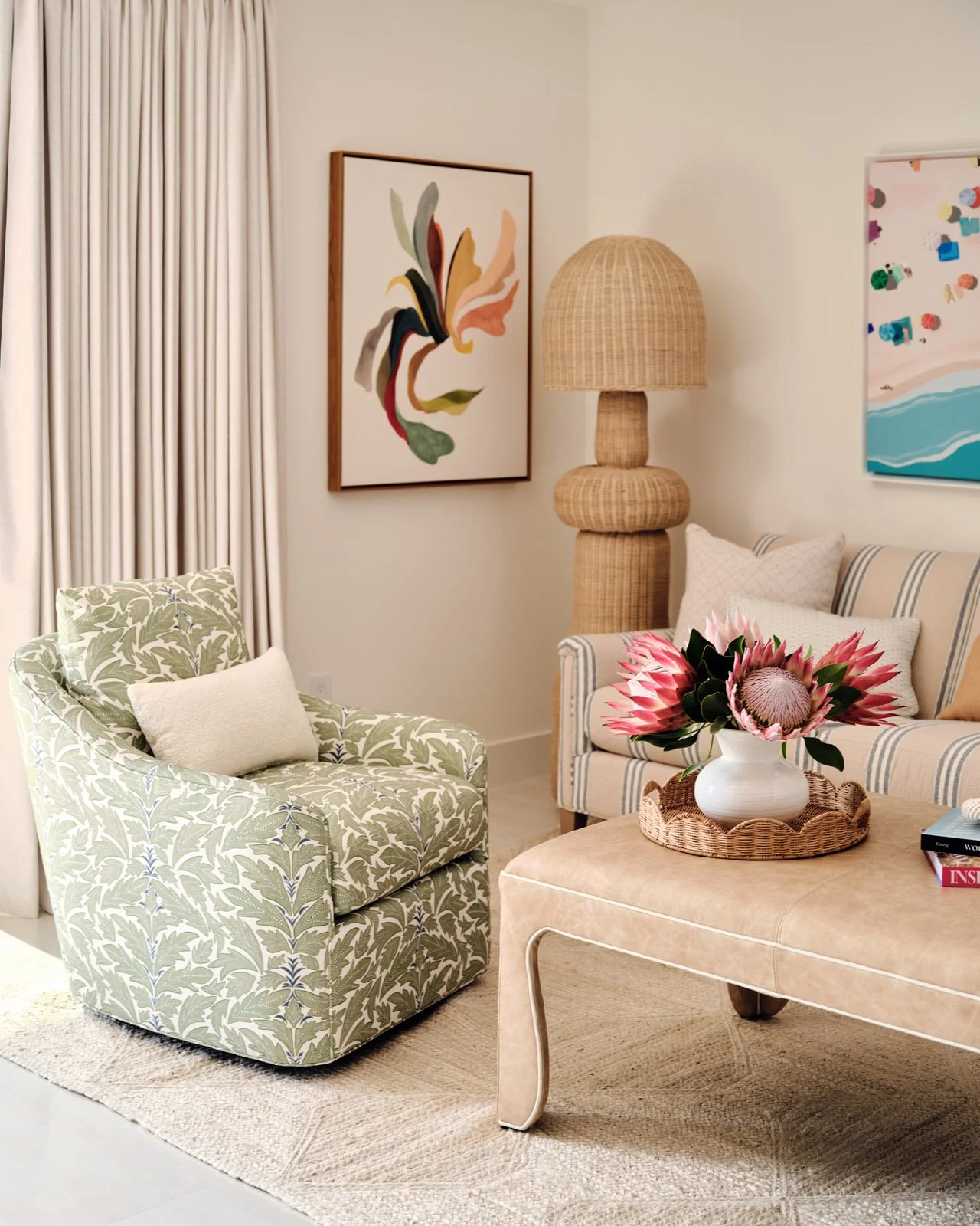 Living room with a patterned armchair, coordinating sofa, a wicker base floor lamp with a woven shade, a framed abstract artwork, a vase of pink flowers on a beige ottoman, and beige curtains.
