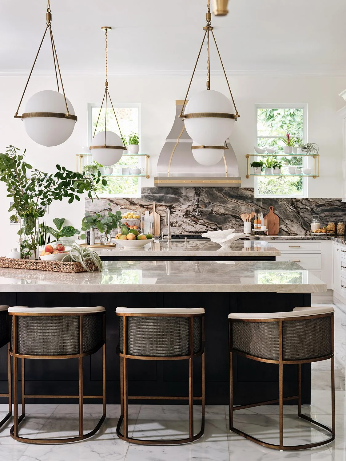 Modern kitchen with white walls, pendant lighting, marble countertops, black island, bar stools, and greenery.