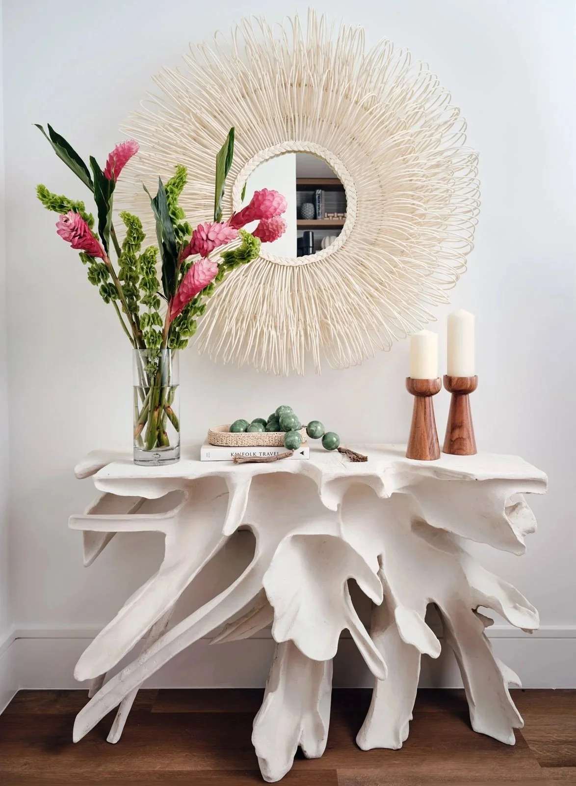 Decorative white driftwood console table with a large glass vase of pink and green flowers, two wooden candlesticks with white candles, a small tray with green decorative balls, and a book titled 'KINFOLK TRAVEL'. A large round mirror with a woven fr