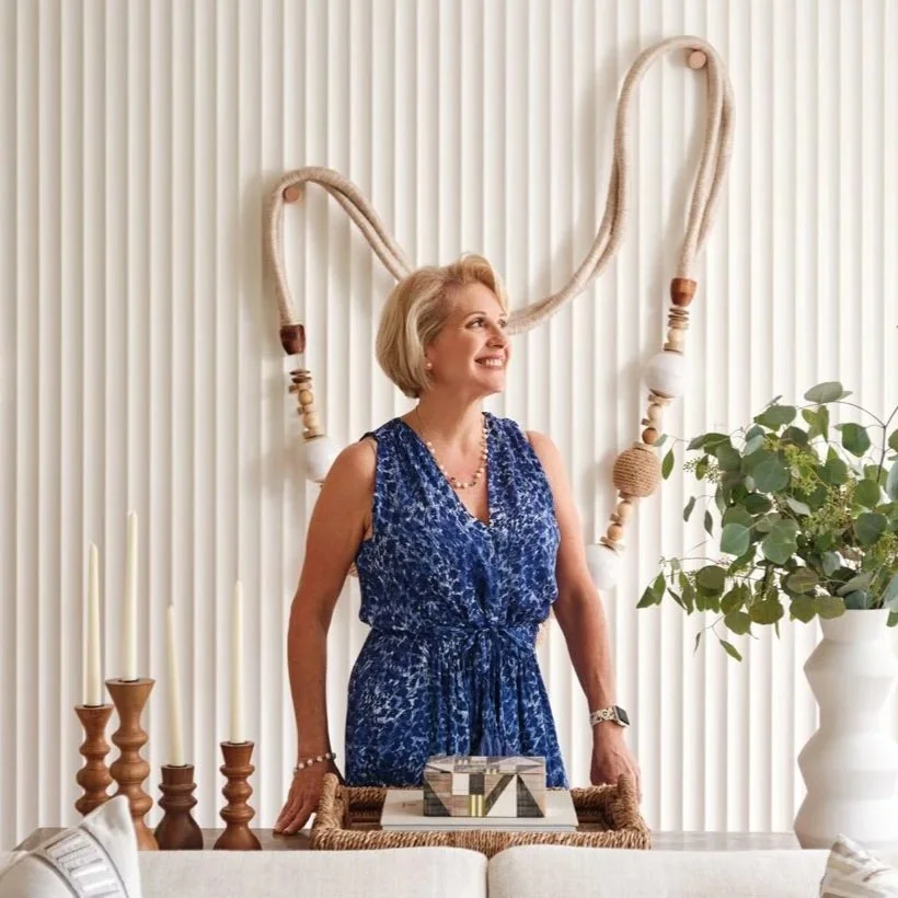 Interior designer Maritza Capiro smiling and standing behind a table and in front of a decorative wall hanging made of large, intertwined rope loops, and to her right is a large white vase with green leafy plants.