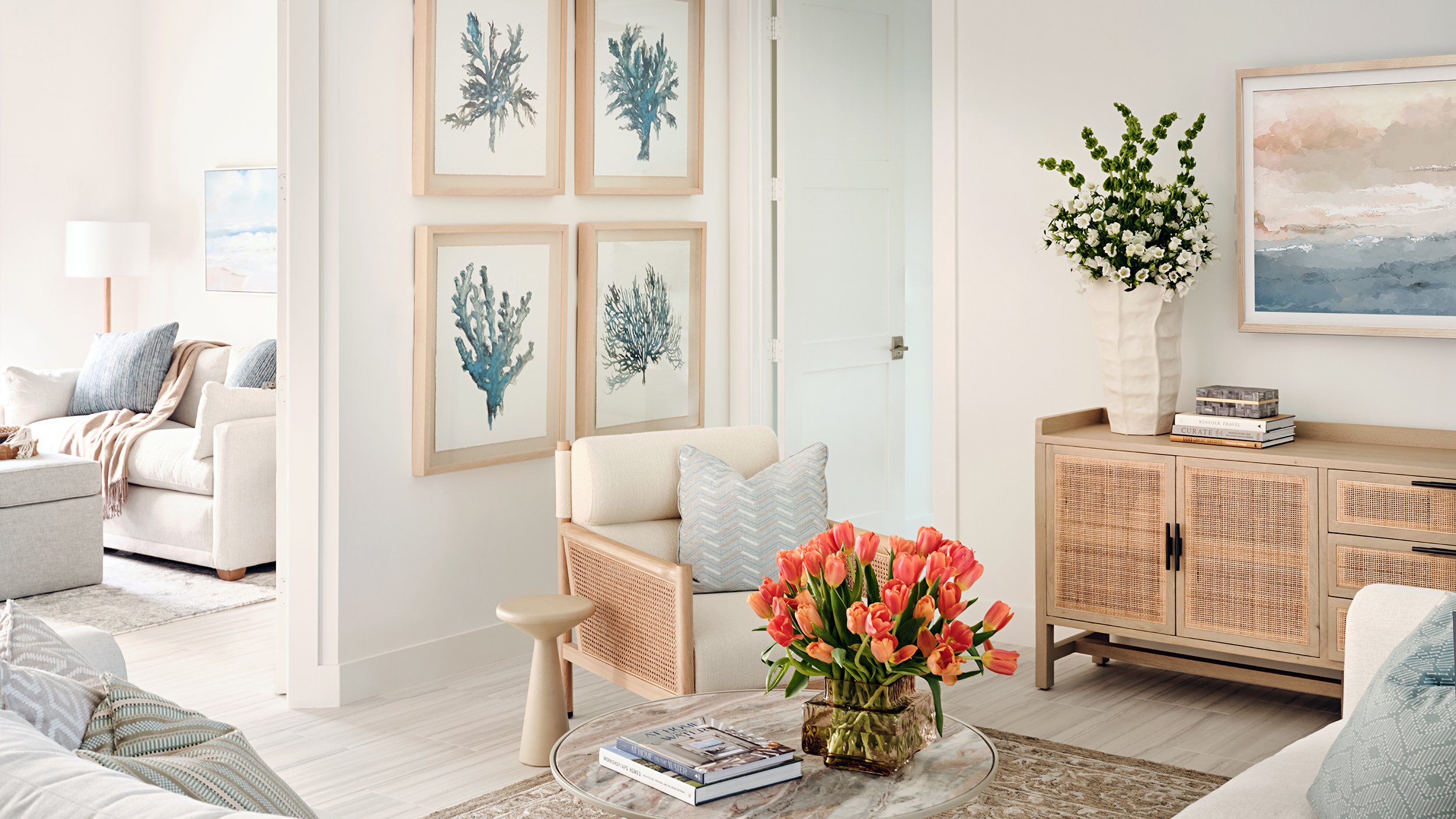 Bright living room with framed coral-inspired artwork on white walls, a beige sofa with pillows, a side table with pink tulips, and a wooden sideboard with a large white vase of flowers, coffee table with books, and an abstract painting.