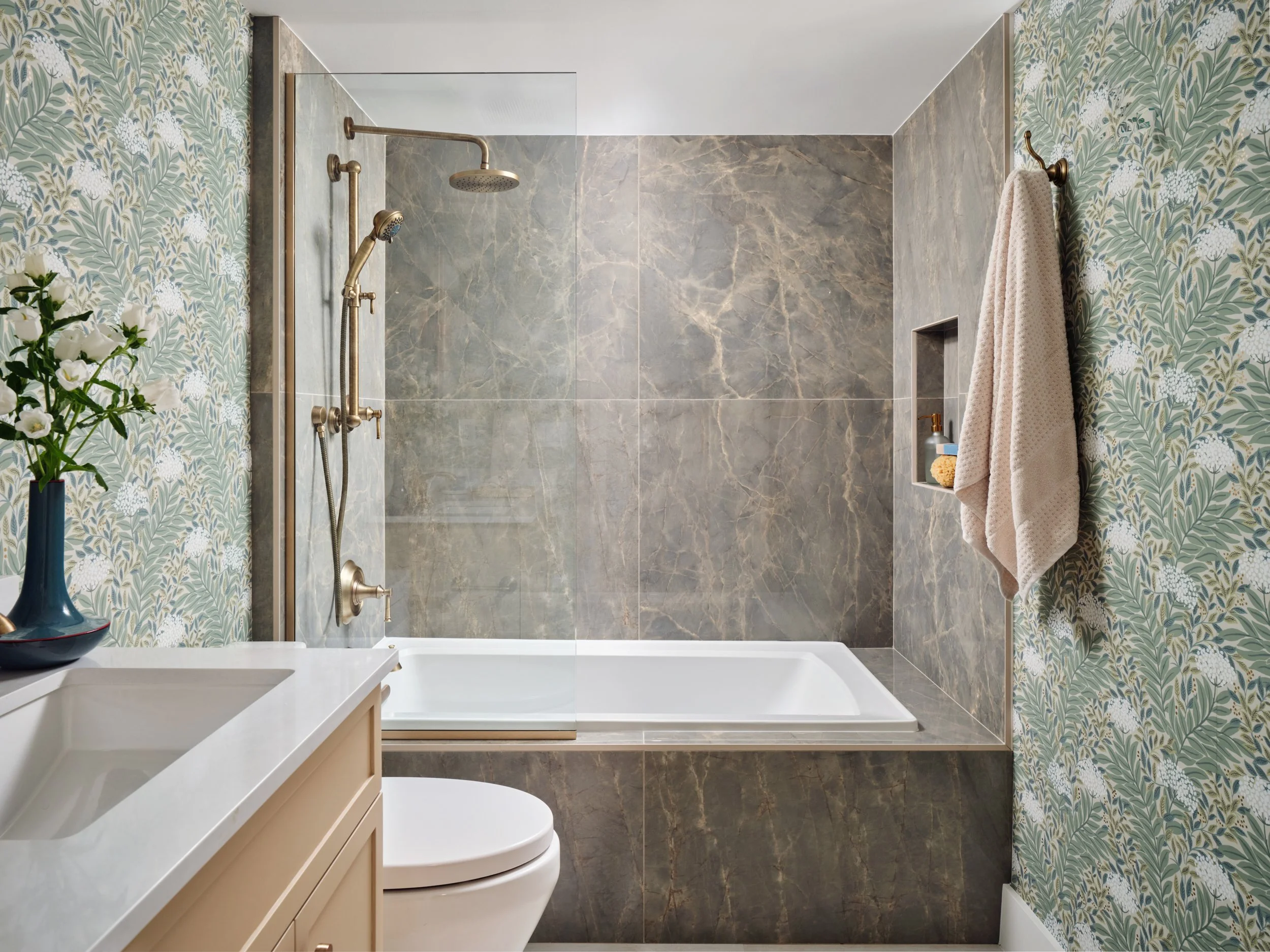 Tub-shower with large veined slabs and a glass panel in a Marco Island condo guest bath with botanical wallpaper
