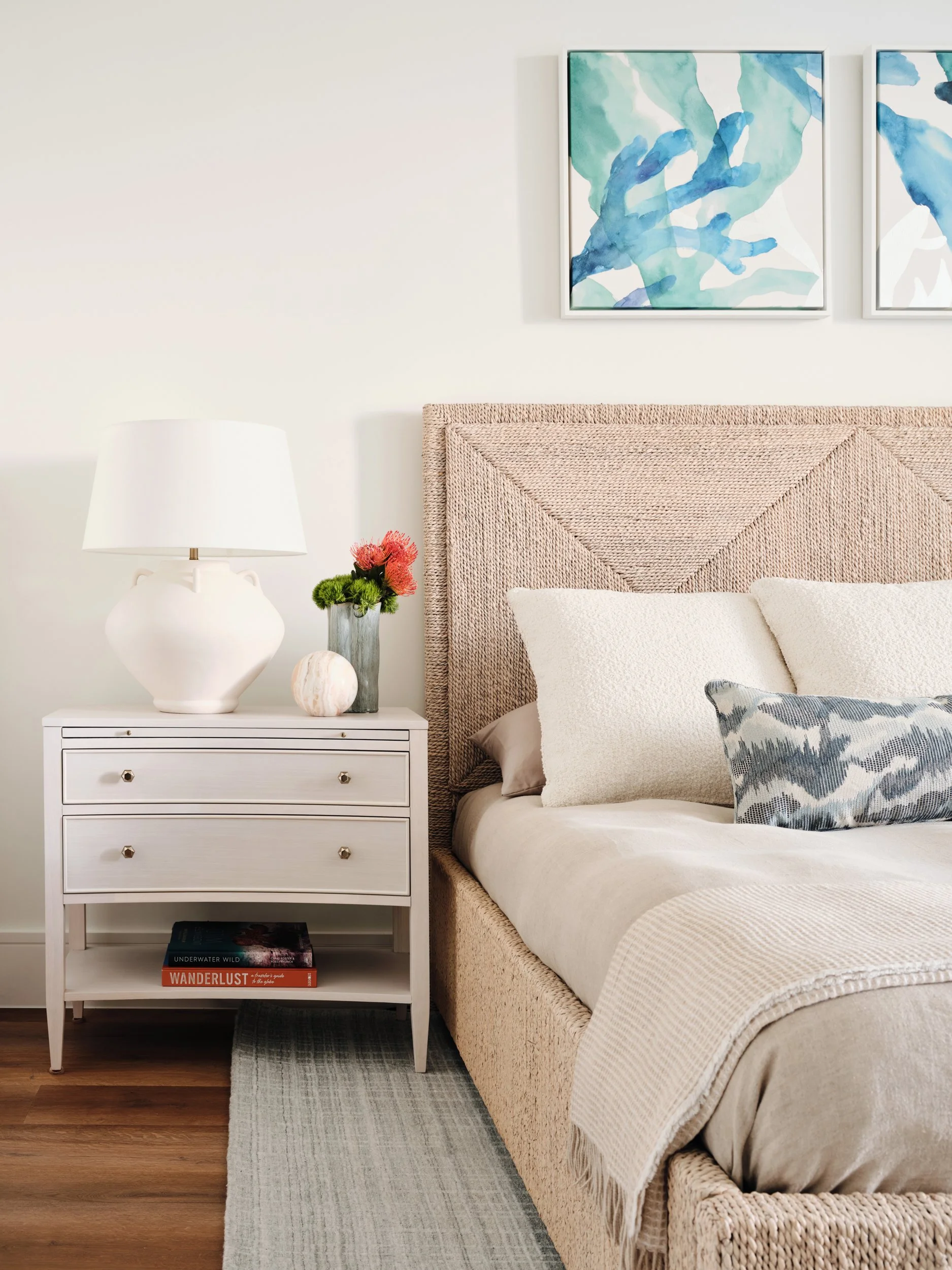 Guest bedroom nightstand with a white ceramic lamp, coral flowers and a marble sphere beside a woven rope bed and blue-green coral watercolor art