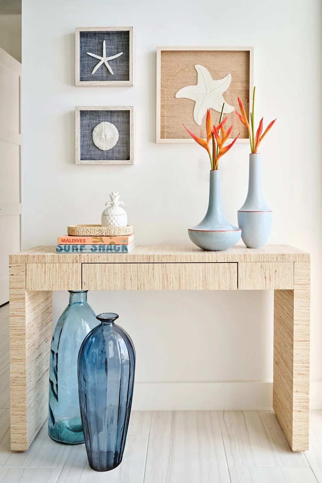 Decorative coastal-themed wall art with starfish and sea urchin shells, a wooden console table with vases of tropical flowers, books, and glass vases in front.