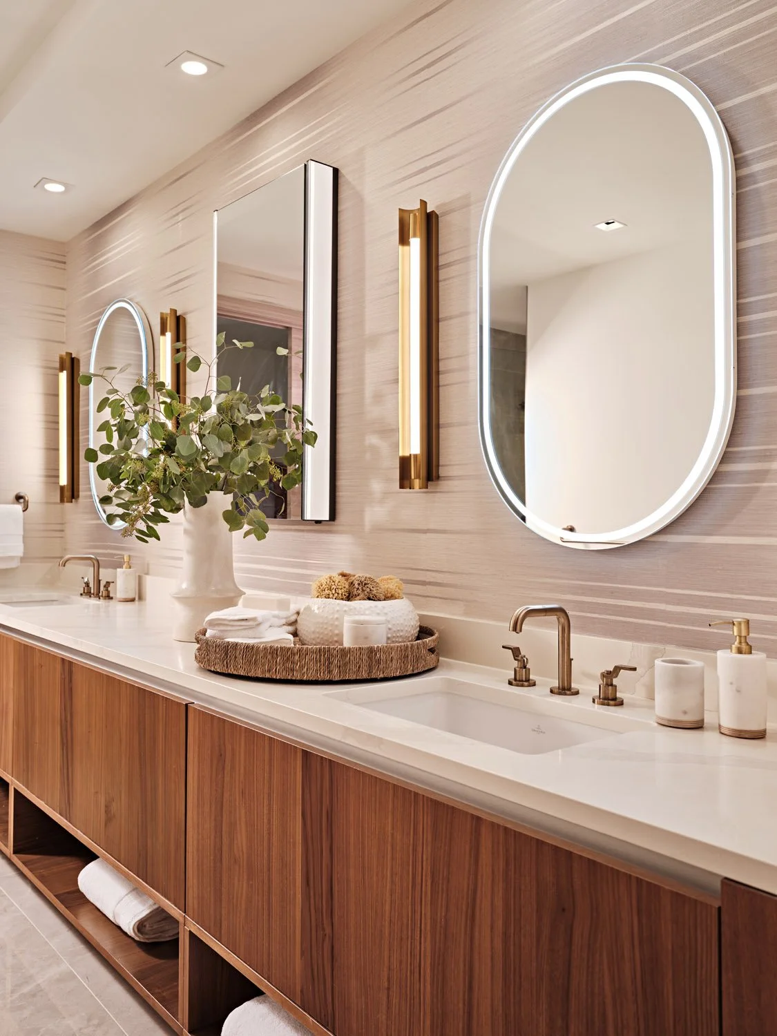 Modern bathroom with a wooden vanity, white countertop, oval mirror with LED lighting, tall vertical mirrors, green leafy plant in a white vase, and towels.