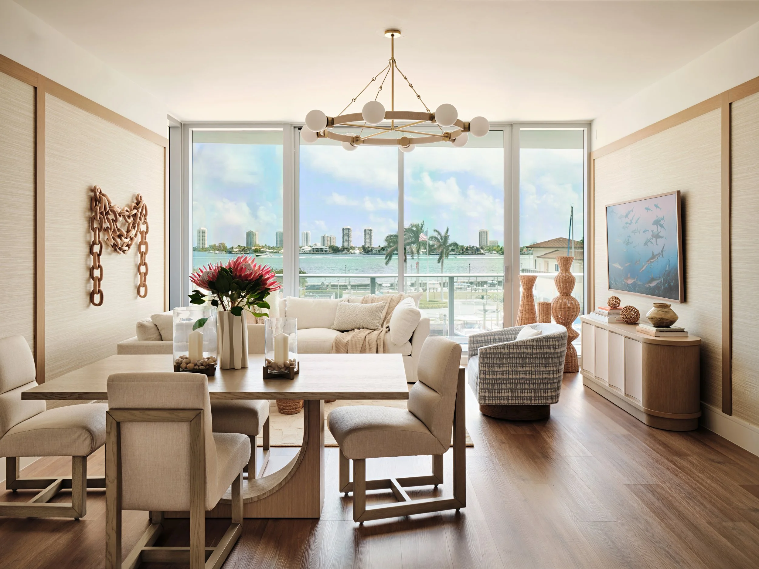 Waterfront condo living and dining room with a ring chandelier, white sectional and grasscloth walls framed in oak