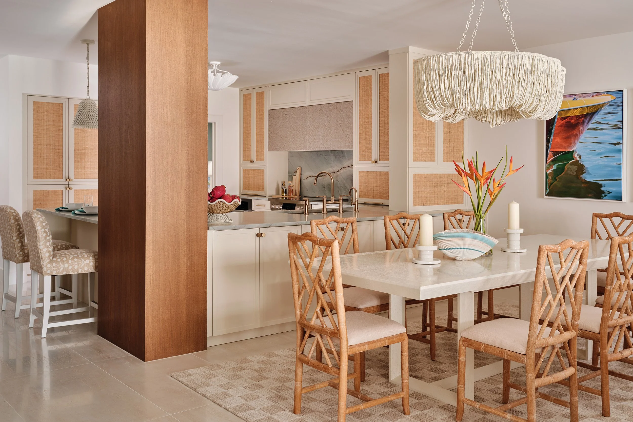 A modern kitchen and dining area with a large dining table, wooden chairs, a white island with bar stools, beige cabinets with woven rattan inserts, a marble backsplash, a large woven chandelier, and a colorful boat painting on the wall.
