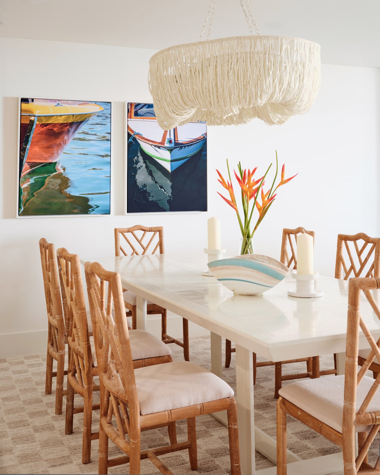 A dining room with a white table surrounded by wooden chairs with white cushions, a vase with orange and red flowers, candles, and two photographs of boats on the wall.