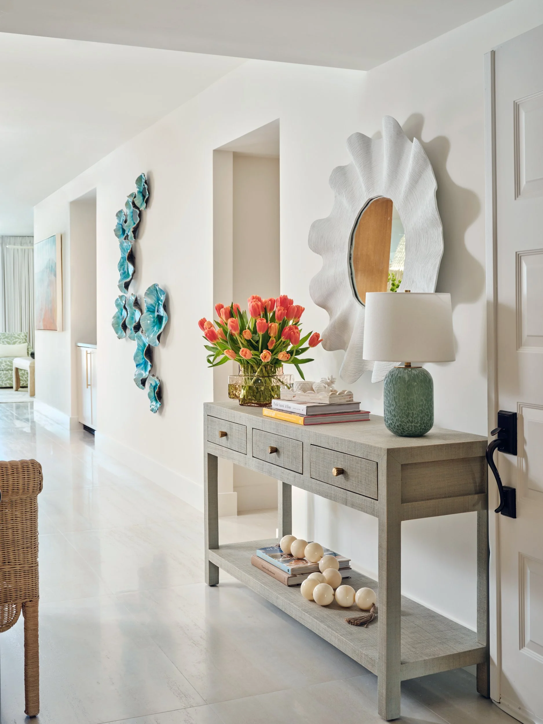 Renovated entry at The Duchess on Marco Island with a woven console, coral tulips, a sculptural white mirror and turquoise wall art