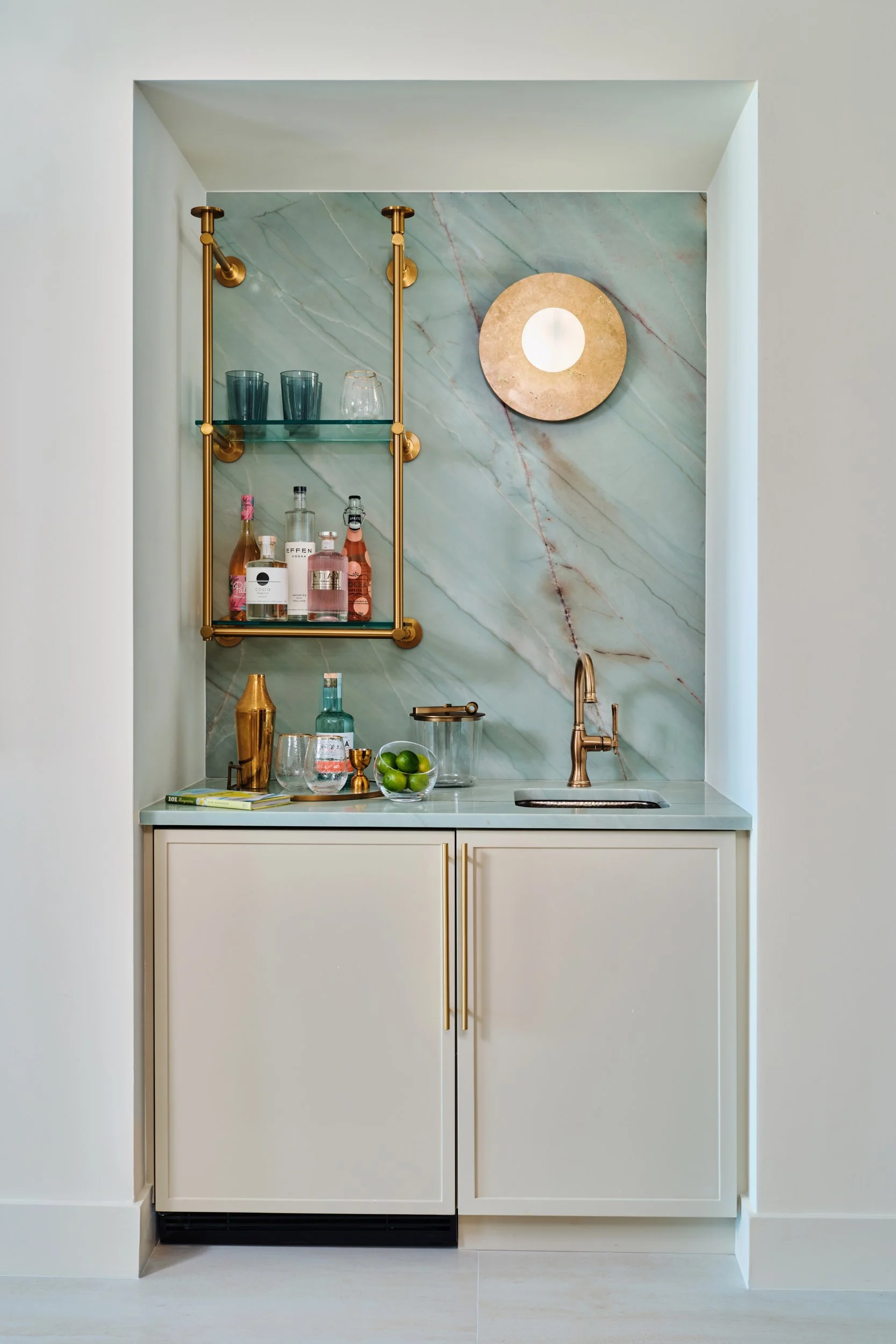 Wet bar at The Duchess on Marco Island with a green quartzite backsplash, brass and glass shelving and a hammered copper sink