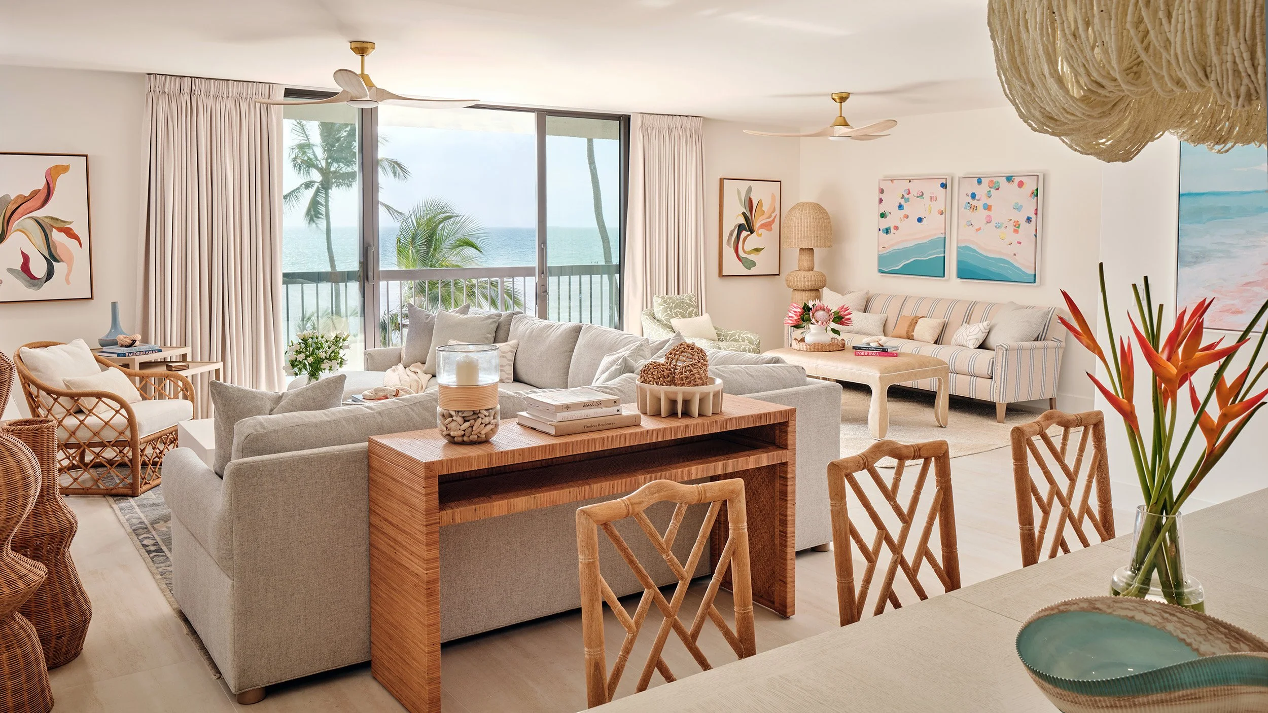 Living room with large sliding glass doors overlooking tropical beach and ocean, decorated with abstract artwork, wicker furniture, plants, wood accents, and light-colored curtains.