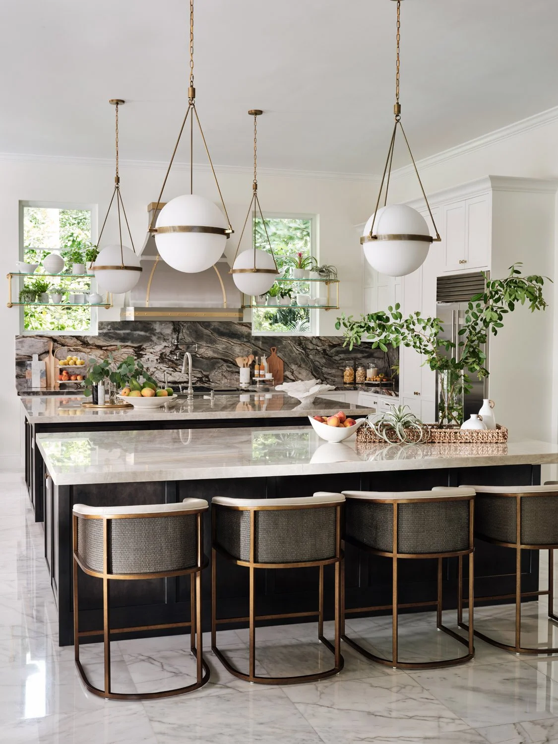 Modern kitchen with marble countertops, large island, bar stools, globe pendant lights, stainless steel appliances, and decorative plants.