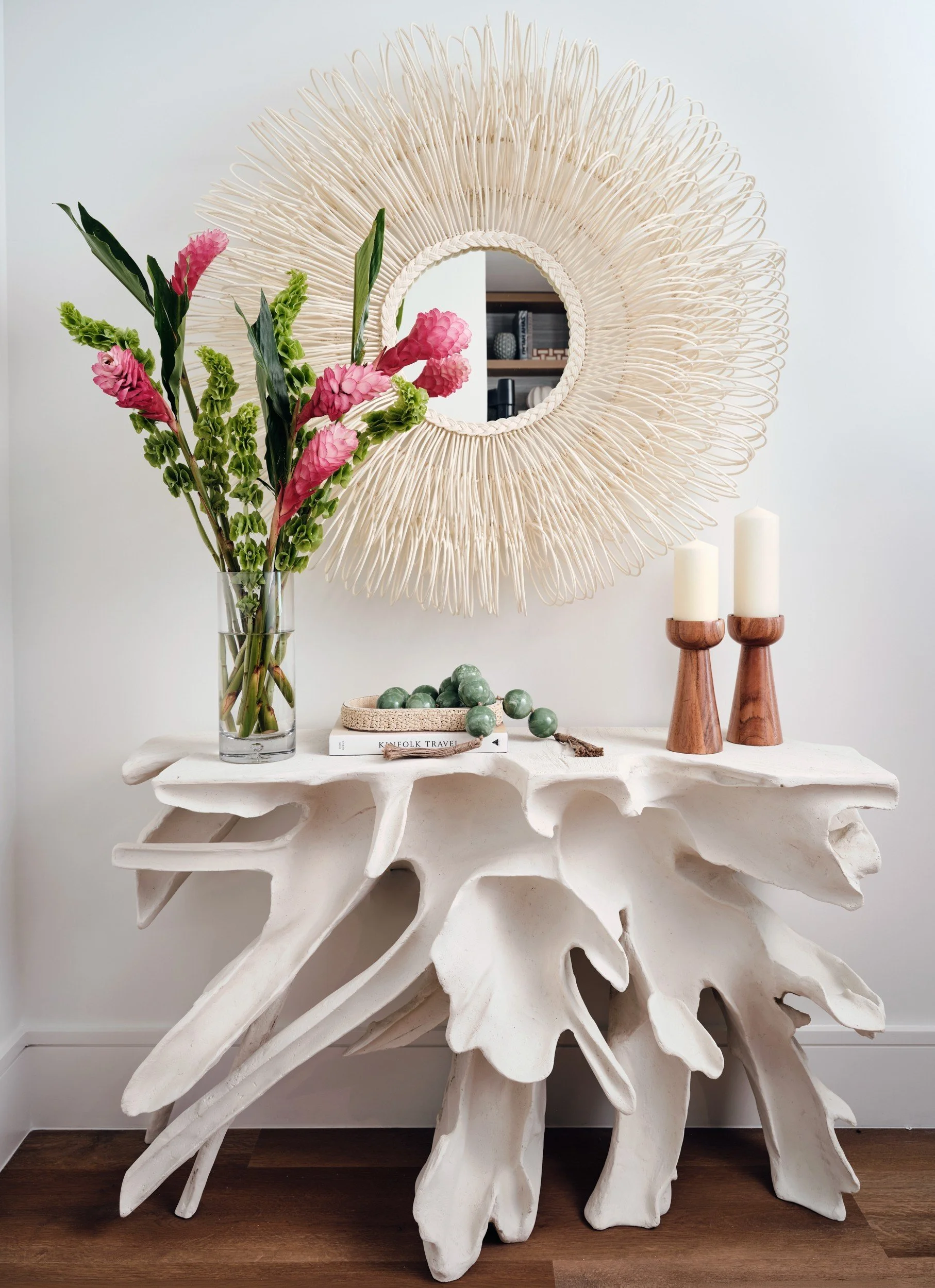 Sculptural white driftwood-style console with a rattan sunburst mirror, pink ginger flowers and wood candleholders in a condo entry