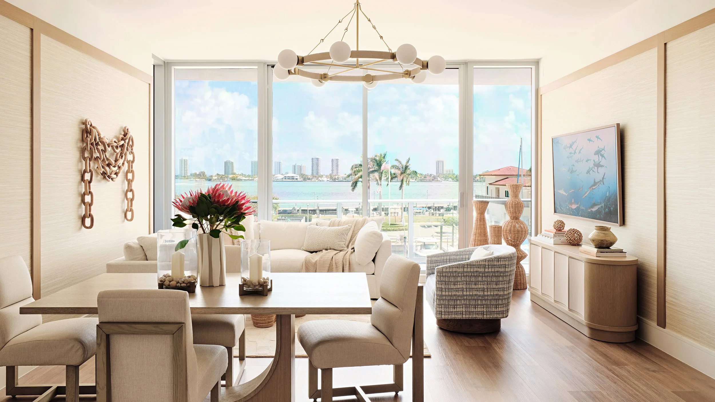Bright living room with large windows overlooking water and city skyline, decorated with neutral furniture, a dining table with candles and floral centerpiece, and coastal-themed artwork and decor.