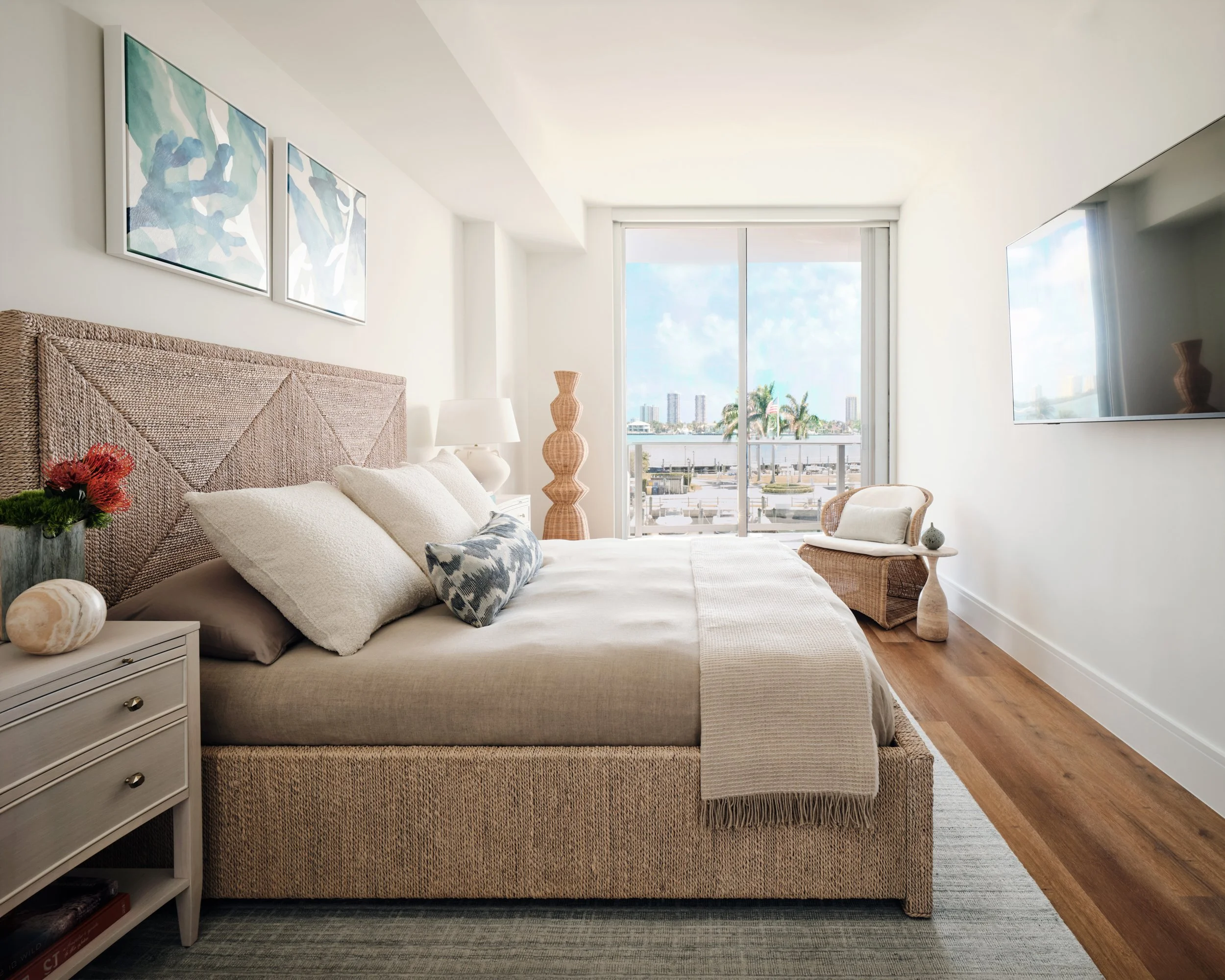 Guest bedroom with a woven rope bed, coral watercolor art and a wicker chair facing the marina