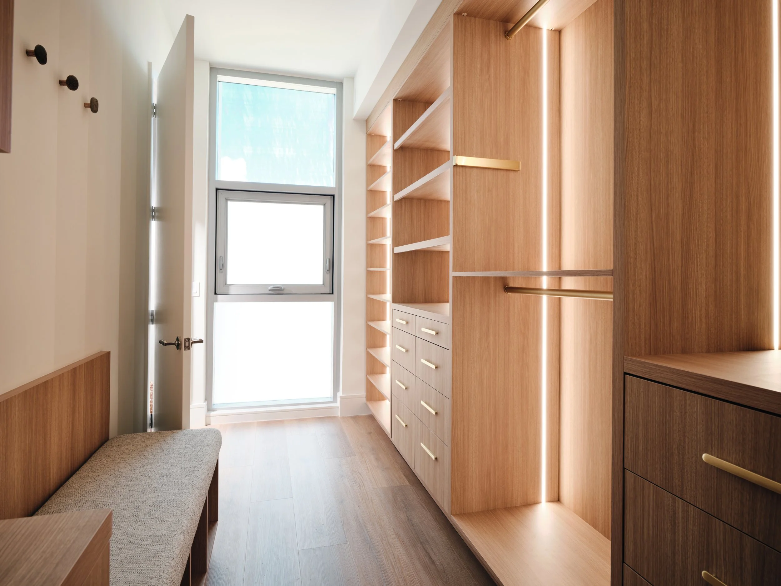 Custom walk-in closet with warm wood shelving, brass hanging rods, built-in lighting, an upholstered bench and wall hooks