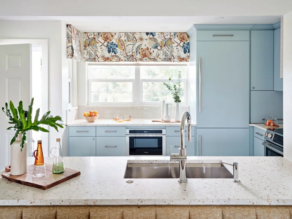 Bright kitchen with light blue cabinets, a window with floral valance, white countertops, a sink with a modern faucet, and plants and decorations including a potted plant, a bowl of fruit, and glass bottles.