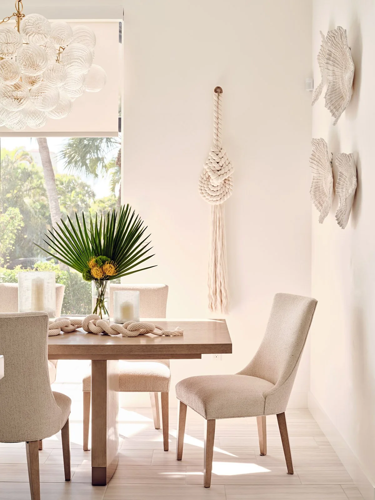 A bright dining room with a wooden table, beige upholstered chairs, a centerpiece with green palm leaves and yellow flowers, candles, and nautical wall decor including a hanging rope knot and shell-shaped wall hangings. Large window with palm trees o