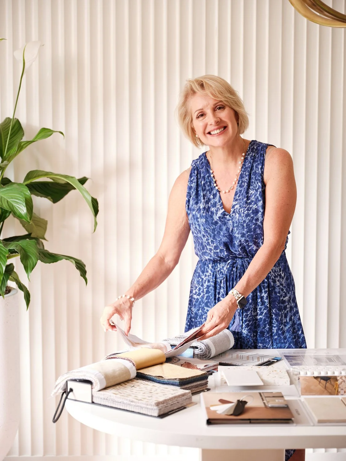 South Florida interior designer Maritza Capiro standing at a table with fabric samples, color swatches, and design materials, in front of a cream-colored textured wall.
