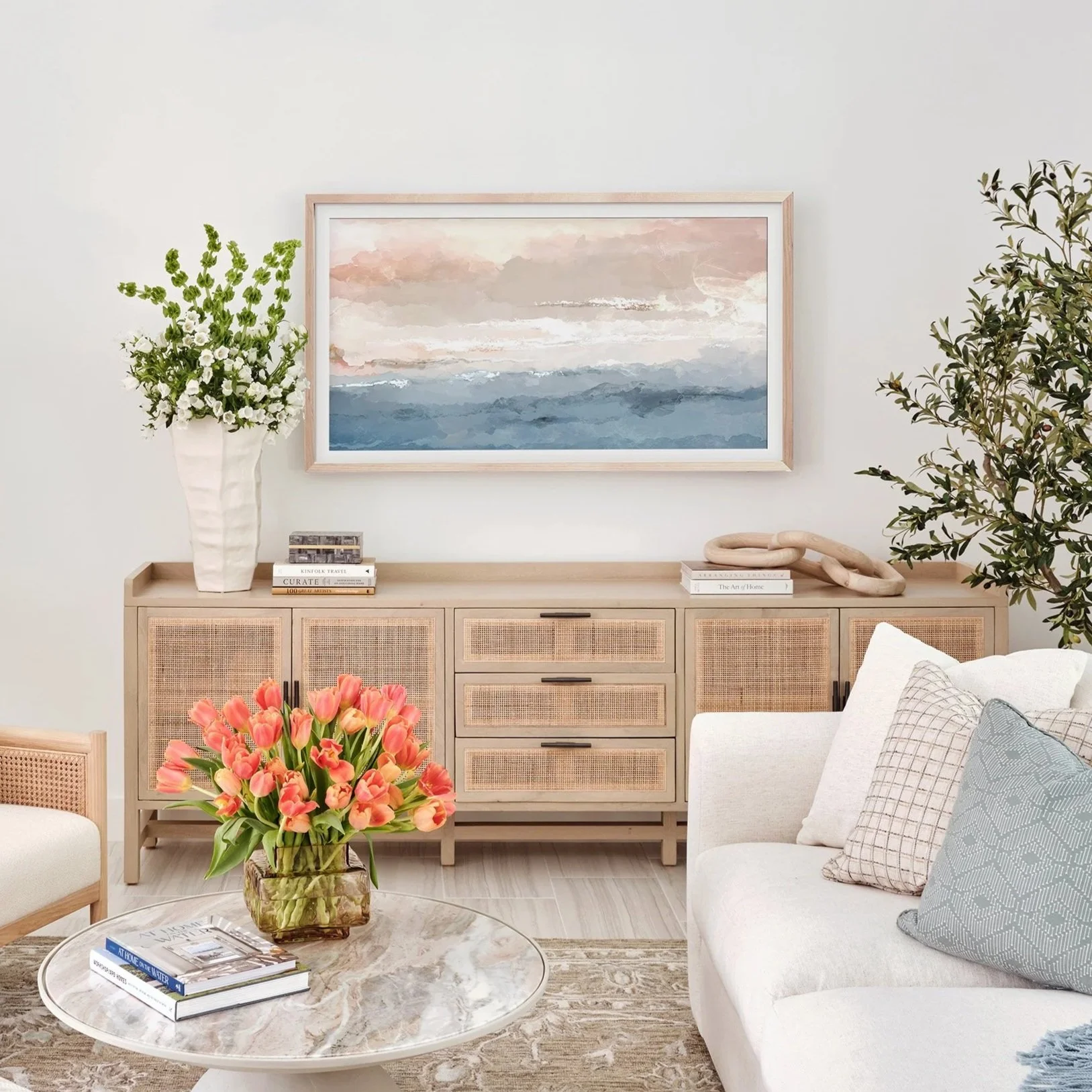 A living room with a beige sideboard, floral paintings, and a white sofa with patterned pillows. A large bouquet of pink tulips in a glass vase is on a round marble coffee table, and a tall white vase with white and green flowers is on the sideboard. There are some books and a decorative wooden chain on the sideboard.