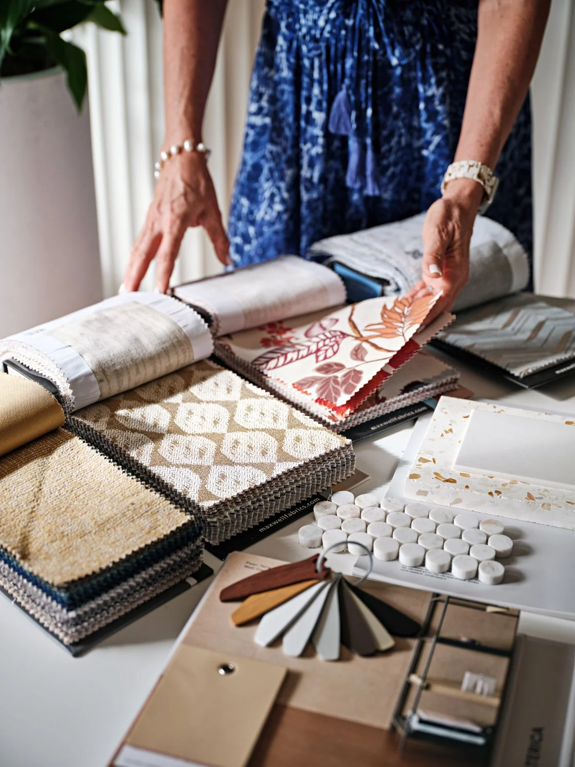 Person in a blue outfit looking at fabric swatches and wallpaper samples arranged on a table for interior design choices.
