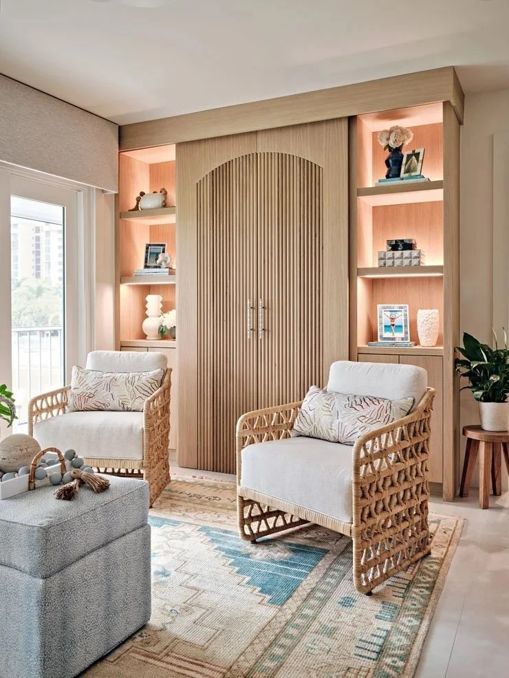 Living room with two rattan armchairs with white cushions, a gray ottoman with toys, a colorful area rug, built-in shelves with decorative items, a wooden sliding door, a potted plant, and a large window.