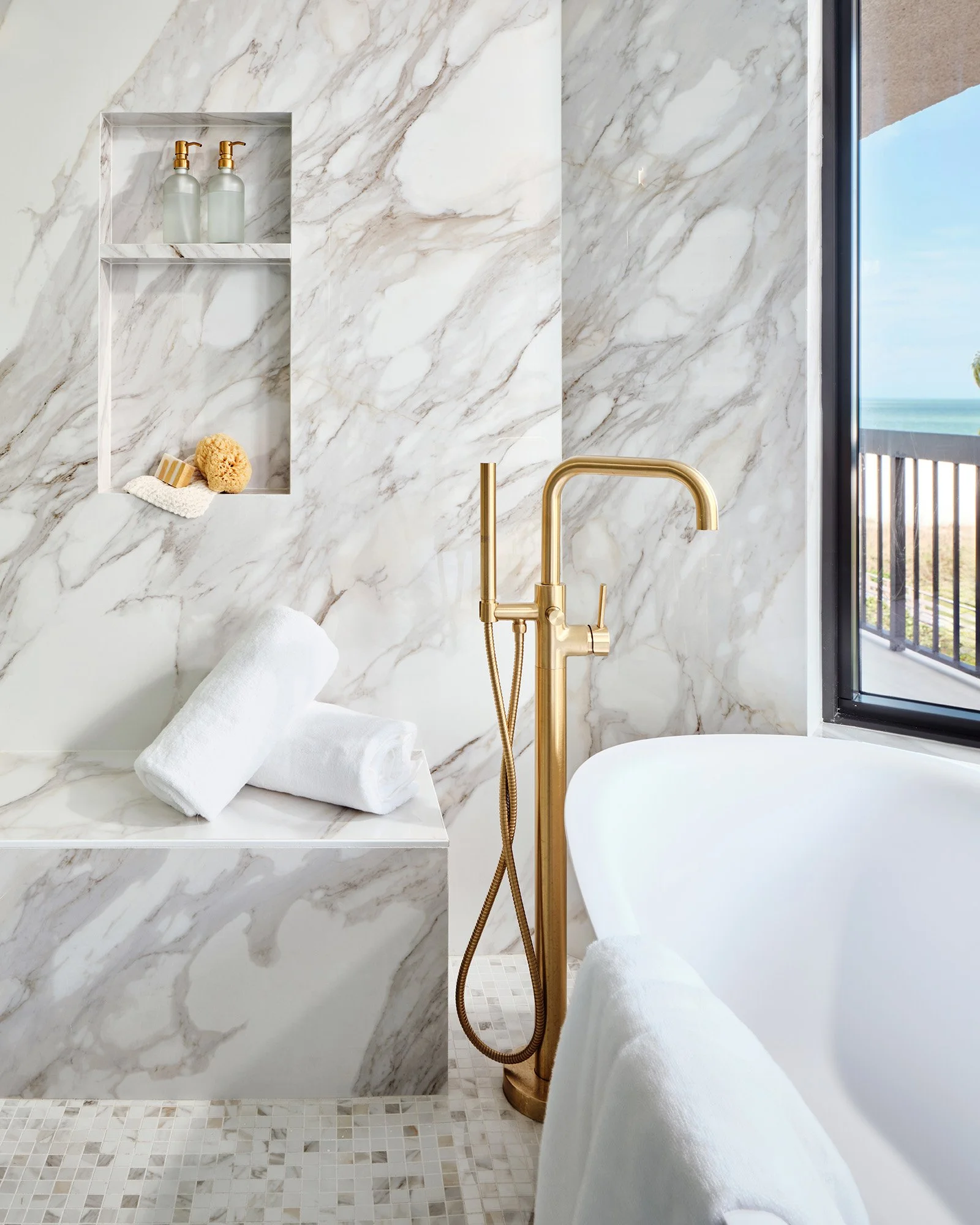 A luxurious bathroom with marble walls and floor, a white bathtub, a gold bathtub faucet and hand shower, a built-in marble shelf with two soap dispensers, a sponge, and a washcloth, with a view of a balcony and ocean outside.