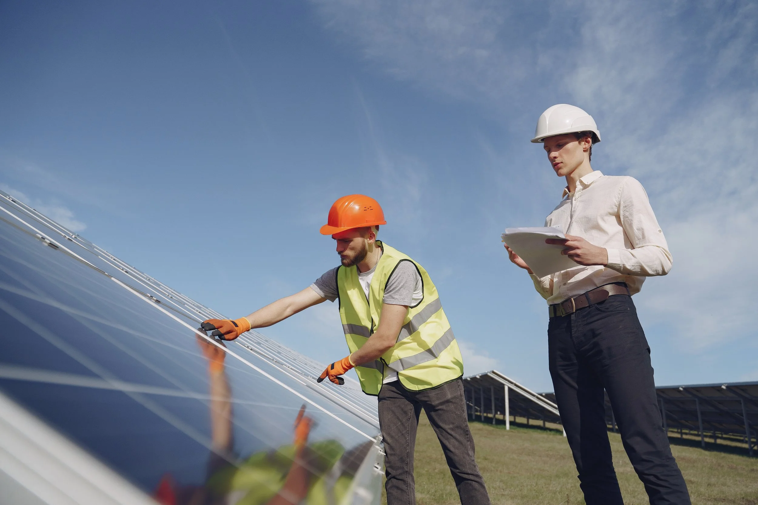Two workers wearing hard hats and safety vests inspecting solar panels outdoors on a sunny day with blue sky.