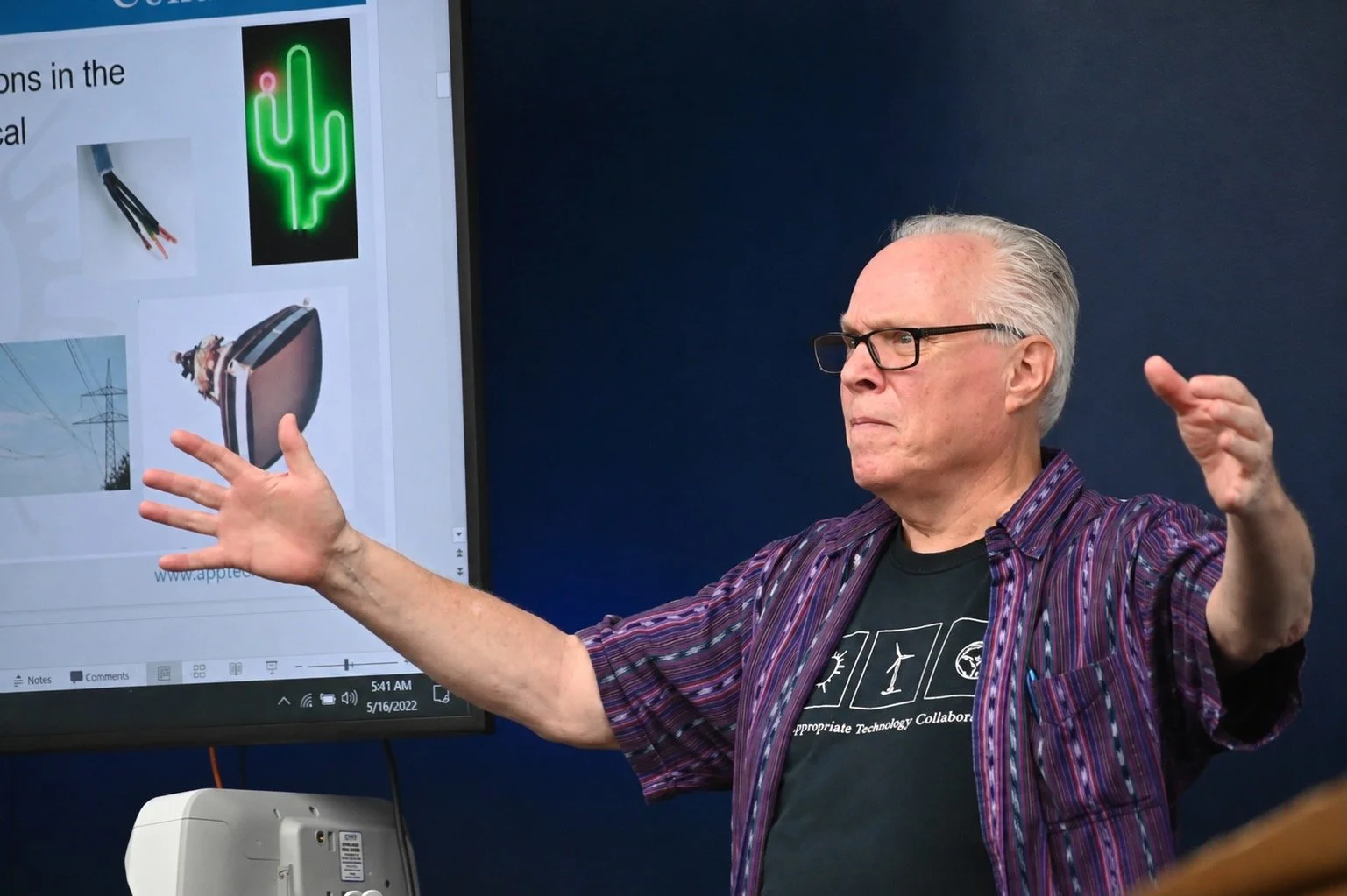 An elderly man with glasses and gray hair, wearing a black t-shirt and a purple plaid shirt, gestures with both hands in front of a large screen displaying images of electrical wires, a neon cactus, and a power transformer, during a presentation or class.