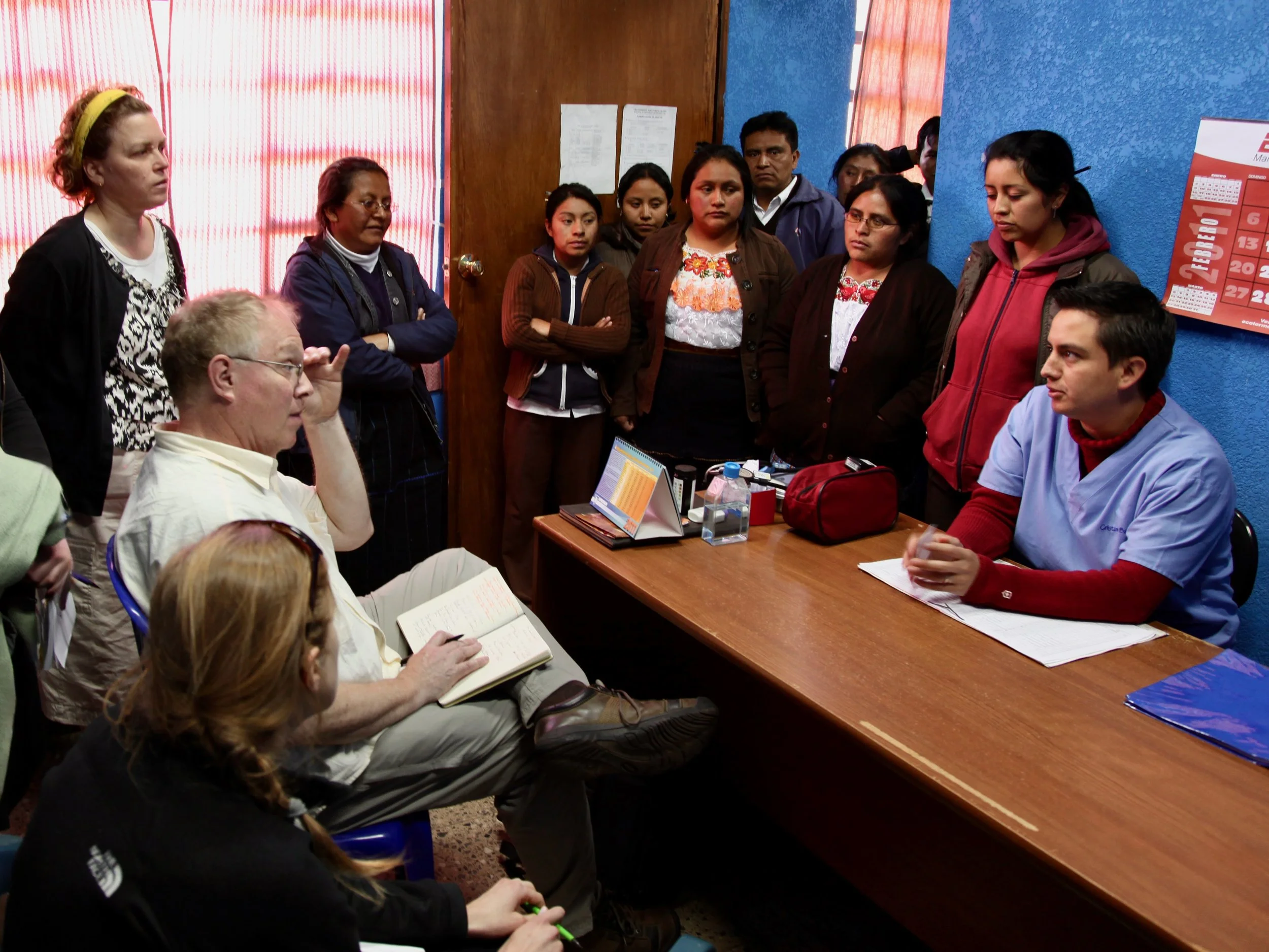 A group of people gathered in a room, listening to a woman speaking. The room has blue walls, a wooden door, and red curtains. Some people are taking notes or looking attentively.