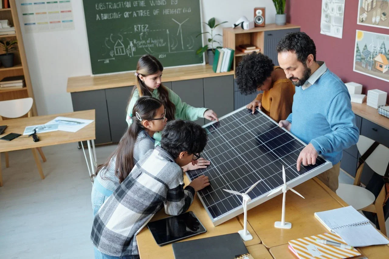 A group of children and an adult examine a large solar panel in a classroom decorated with educational posters and wind turbines models.