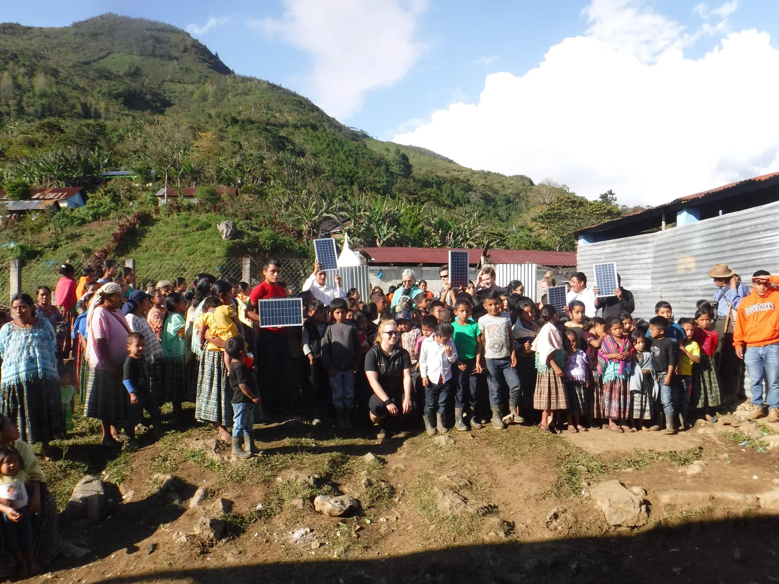A large group of people gathered outdoors in a rural area with mountainous terrain, some holding solar panels.