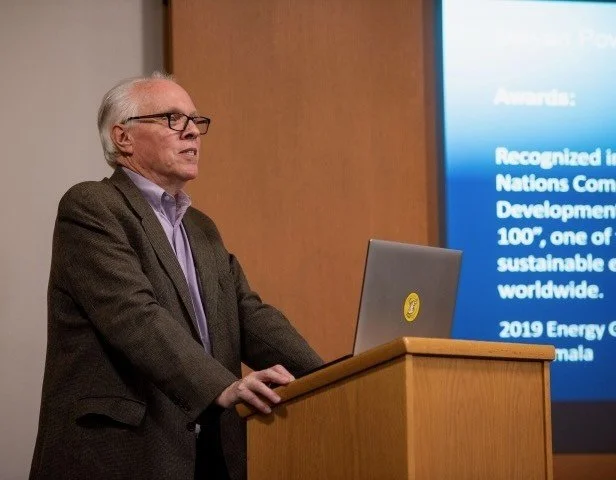 An older man with glasses and grey hair is standing at a wooden podium, giving a presentation. A laptop with a yellow sticker is on the podium and a large screen displays blue text in the background.