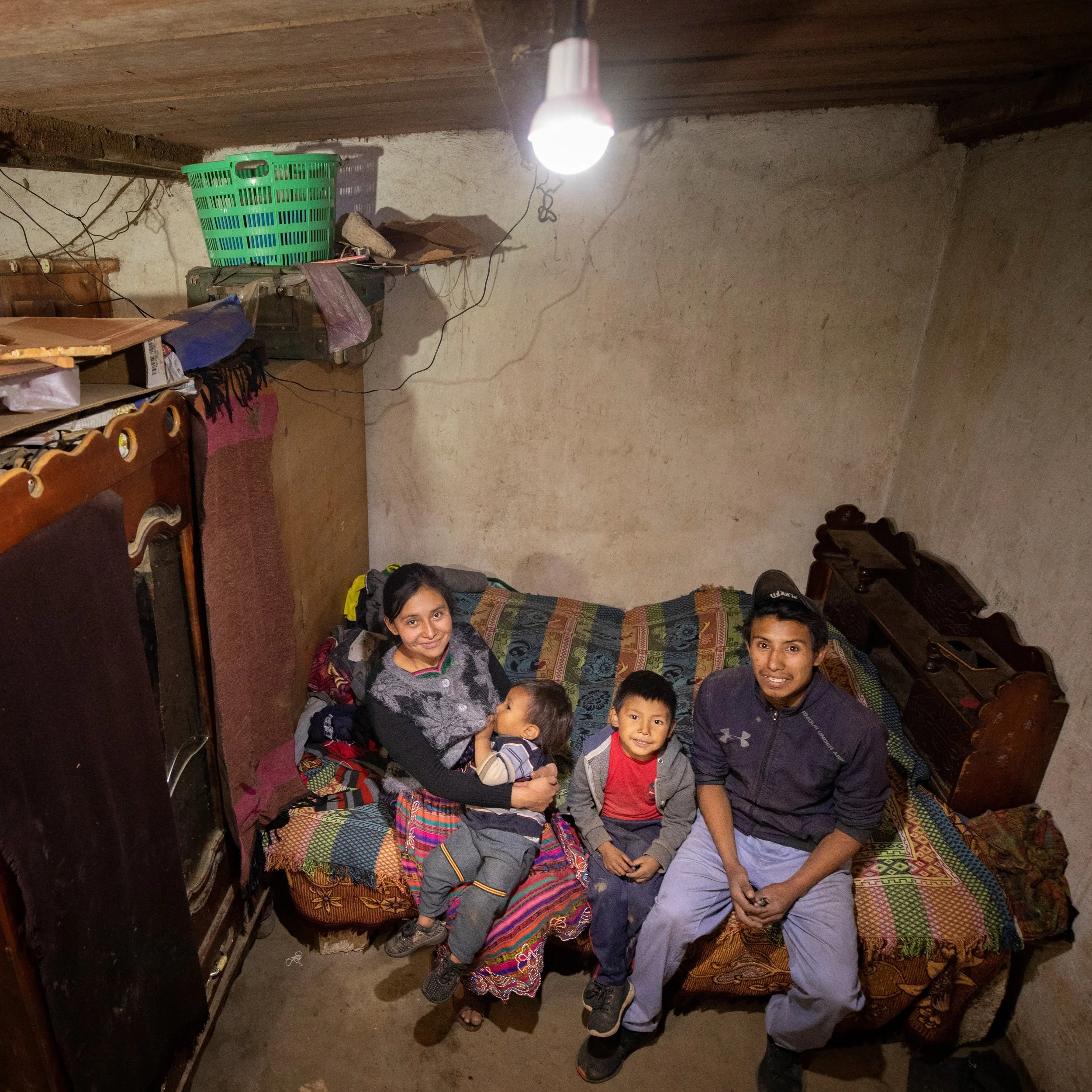 A group of people, including children and an adult, sitting on a patterned bed inside a modest room with beige walls, with a hanging light bulb from the ceiling. There is a green plastic basket and various objects on the shelf behind them.