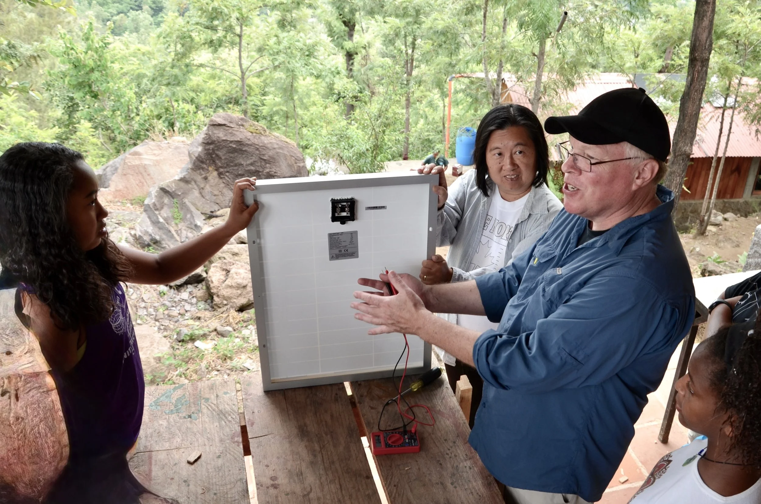 Group of people outdoors examining a solar panel connected to a multimeter, with trees and rocks in the background.